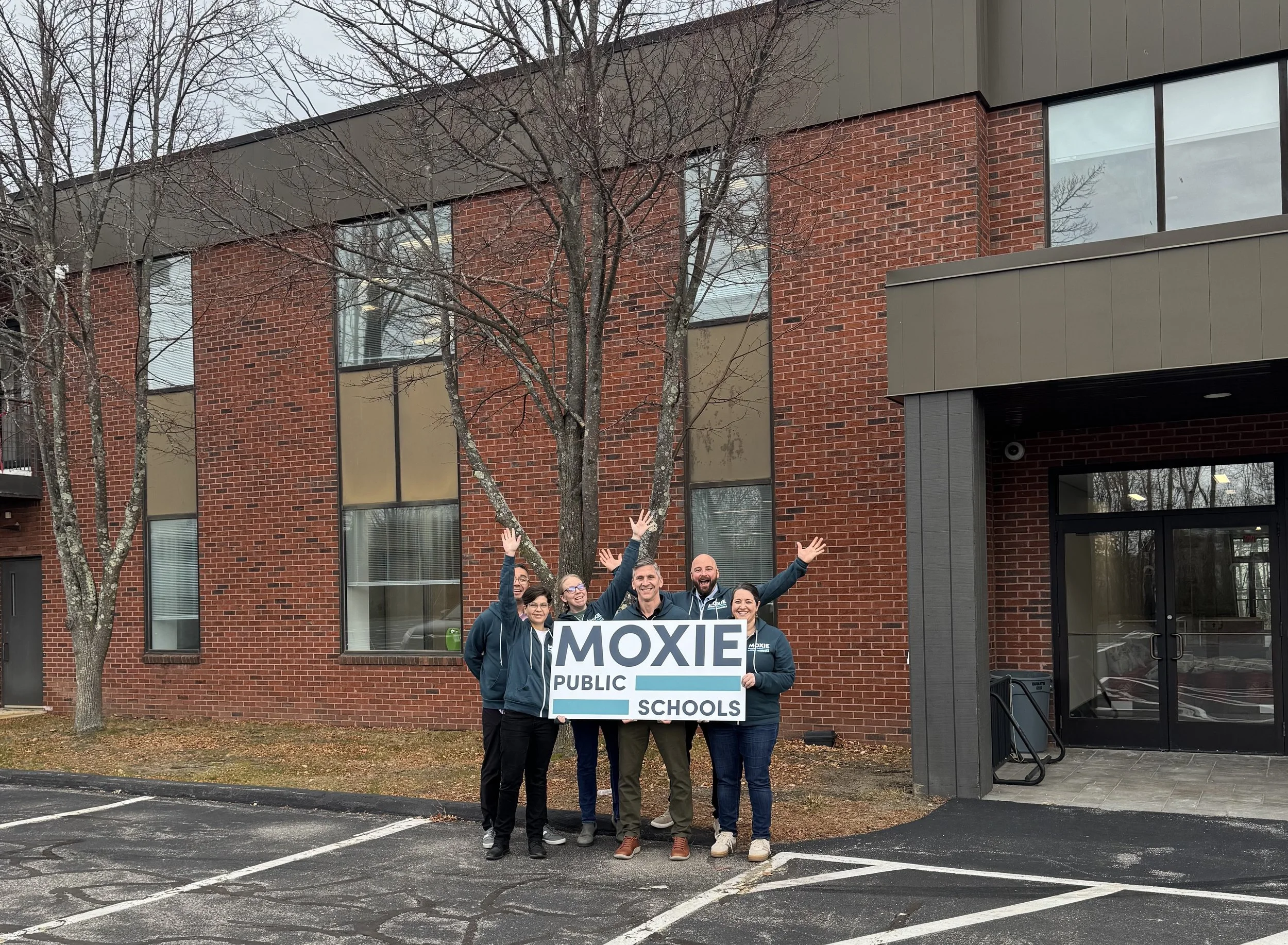 Group of people smiling and holding a sign that says 'MOXIE PUBLIC SCHOOLS' outside a brick building with leafless trees and parking lot.