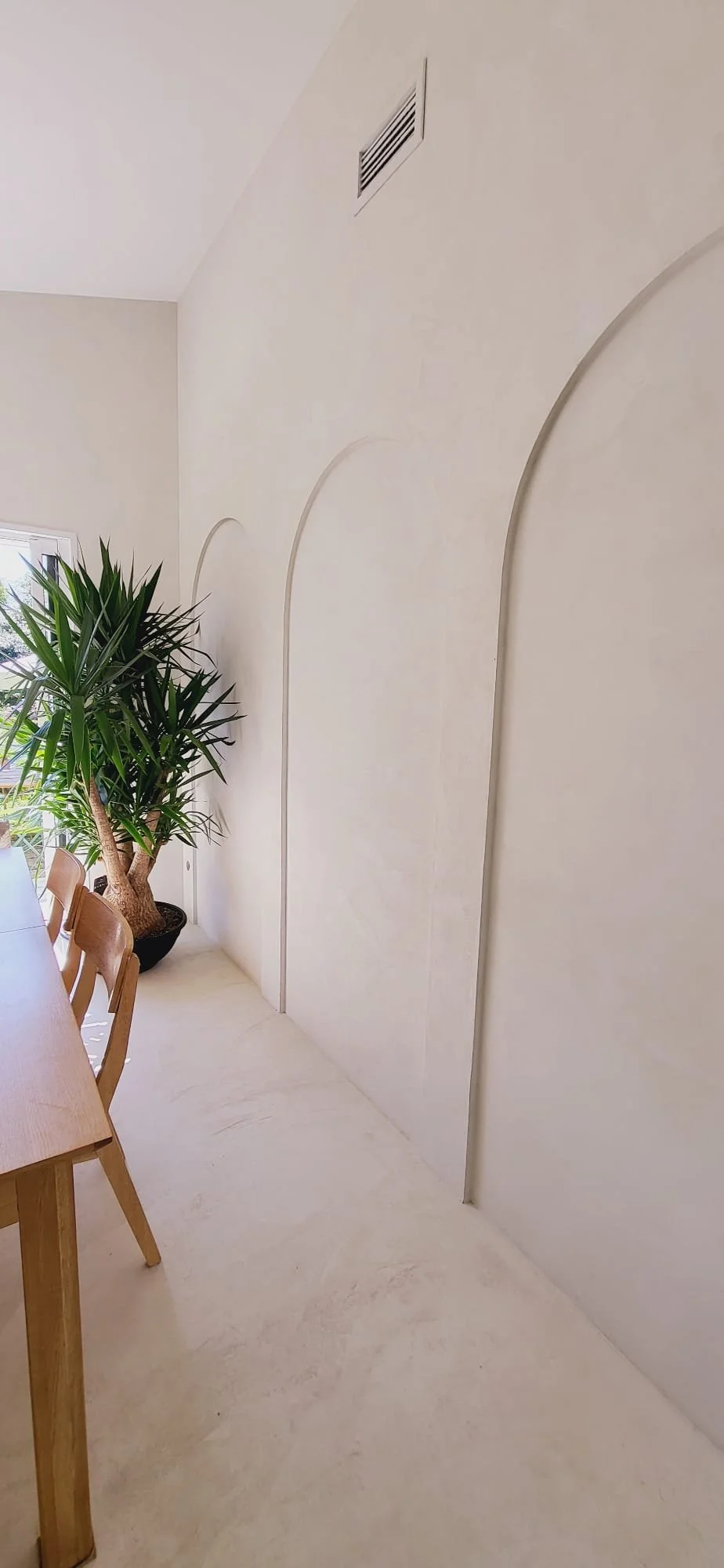 Un mur beige avec des motifs en arc, une plante verte en pot, une fenêtre, et une table en bois avec des chaises. Architecture d'intérieur. Aménagement d'intérieur. Béton ciré, arches. 