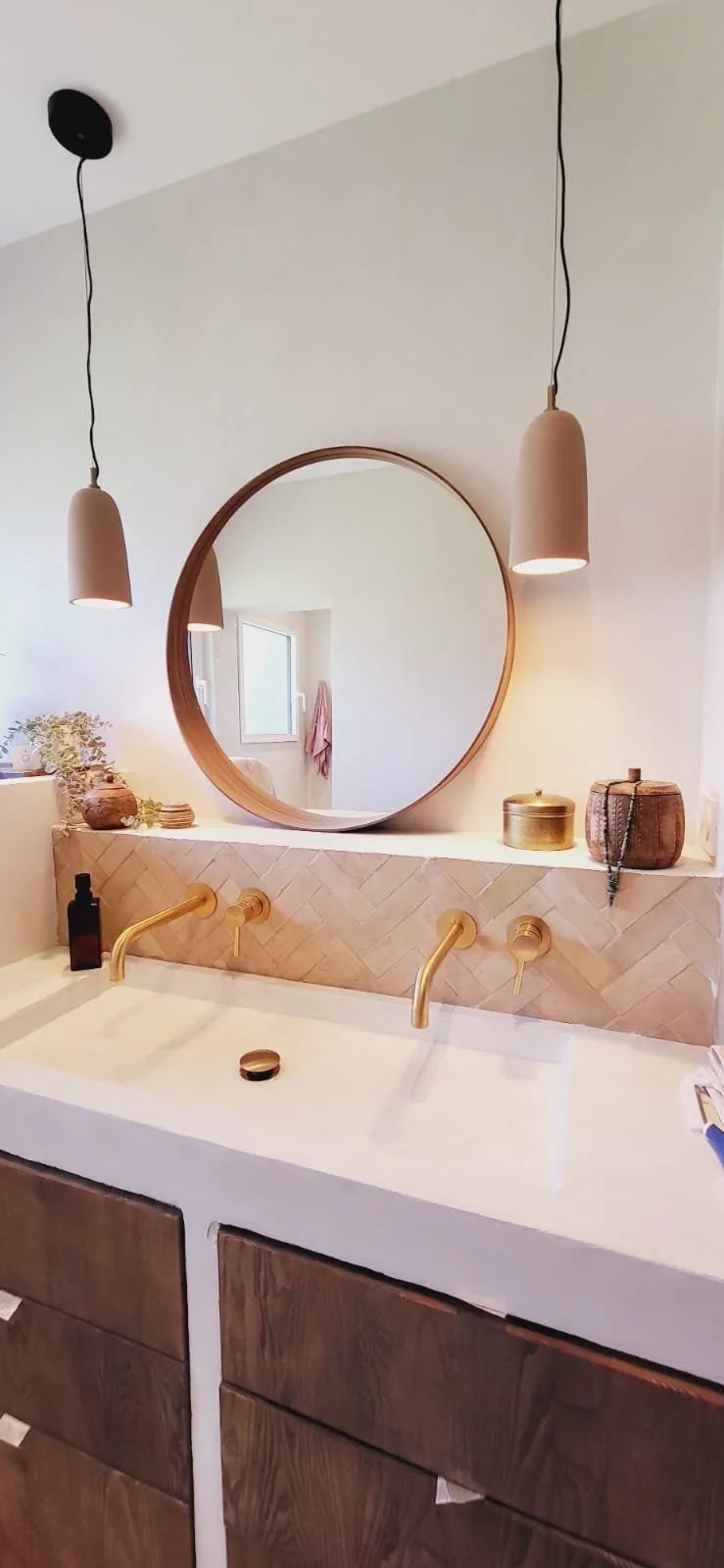 Lavabo avec deux robinets dorés, miroir rond en bois, luminaires suspendus en beige, décoration avec bocaux en rotin, plantes et produits de toilette, mobilier en bois. Architecture d'intérieur. Aménagement d'intérieur. Béton ciré. Bois. 
