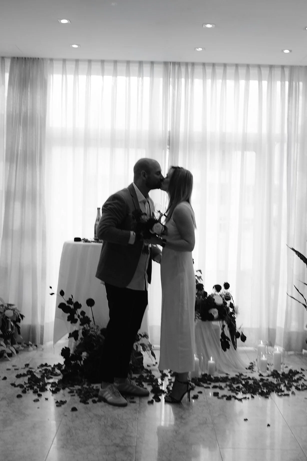 A black-and-white photo of a couple kissing at their surprise proposal in Berlin, holding a bouquet, with candles and flower arrangements on the floor and a curtain backdrop.