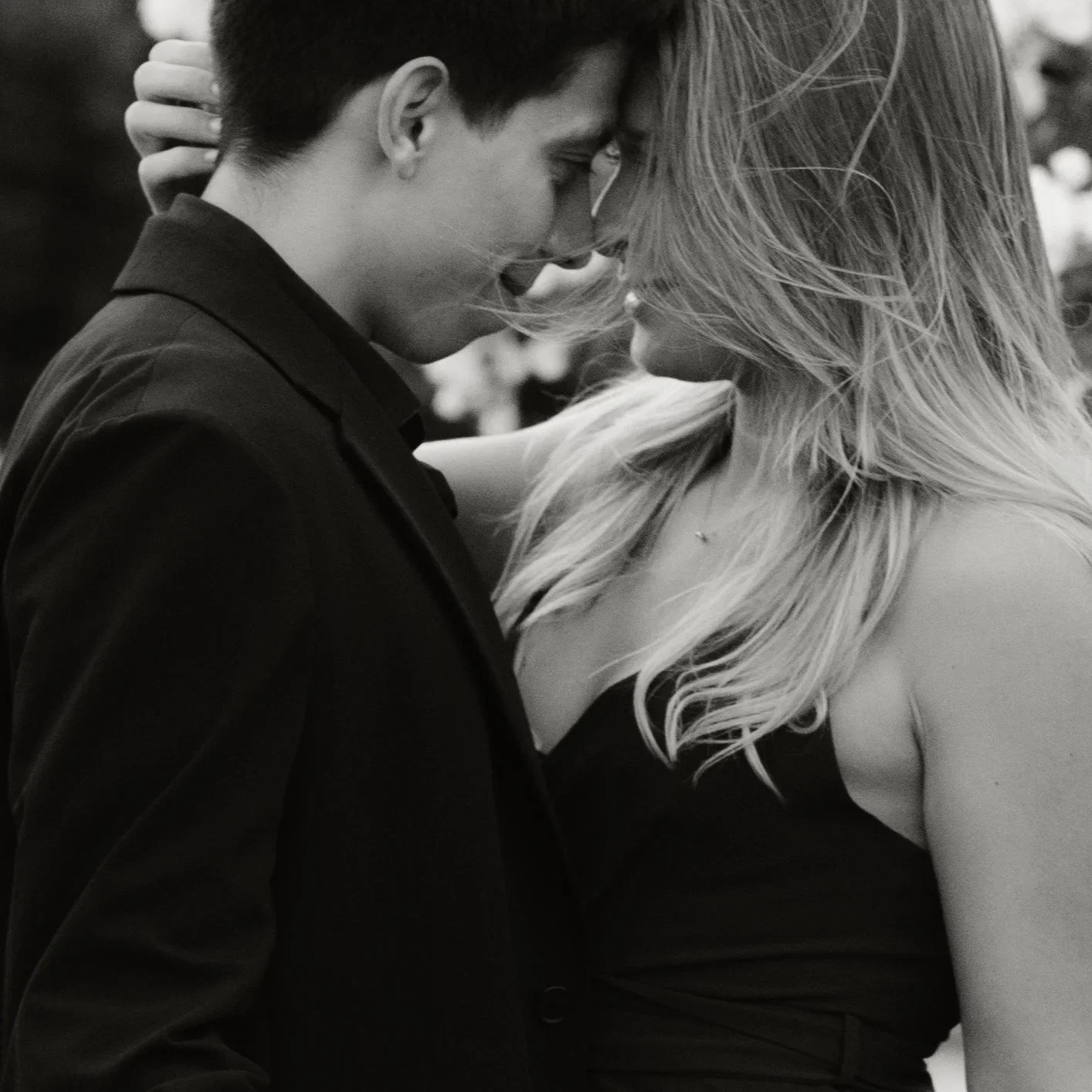 A smiling couple outdoors, with the man wearing a black suit and the woman in a red dress, against a backdrop of greenery and white flowers.