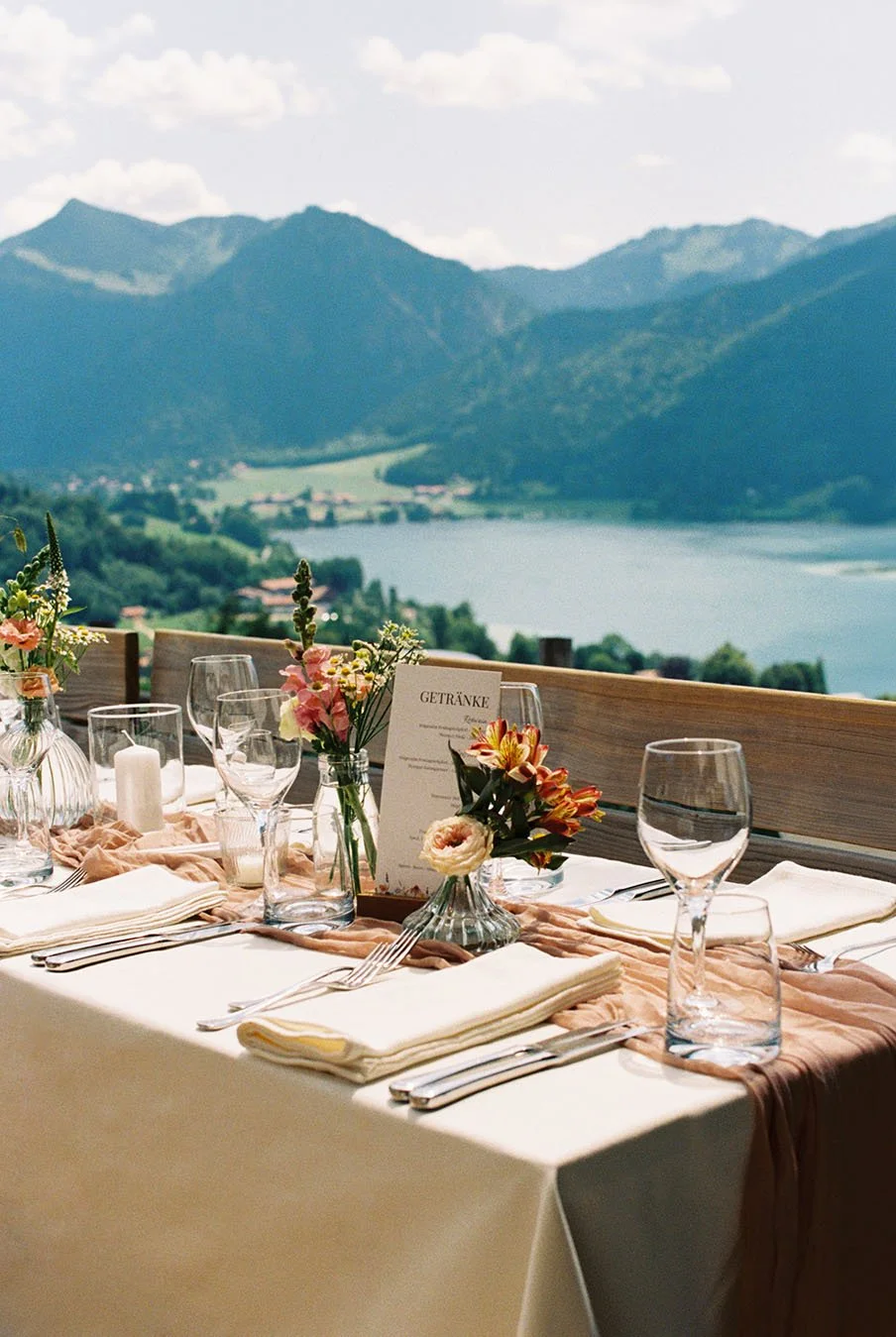 Wedding reception venue with floral decorations, glasses and cutleries overlooking a beautiful lake and surrounding mountains in Germany.