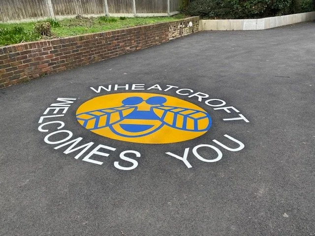 A wheelchair ramp with a painted logo of a smiling face wearing glasses and a yellow circle behind it, with the text 'WHEATCROFT WEE COMES YOU' around the logo.
