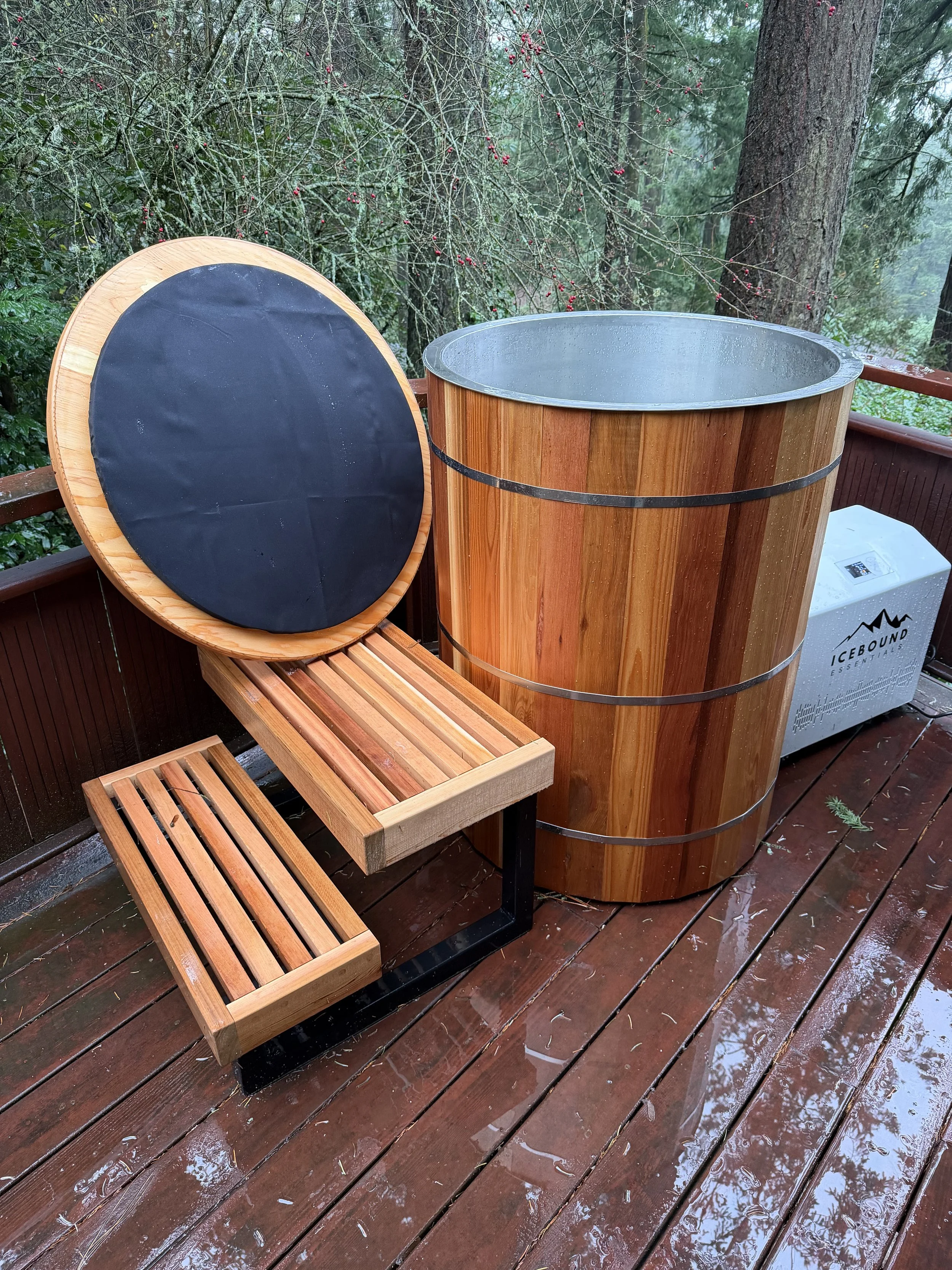 A wooden outdoor hot tub or soaking tub on a wet wooden deck, with a matching wooden bench and a round adjustable stool nearby, surrounded by a lush forest with trees and shrubs.