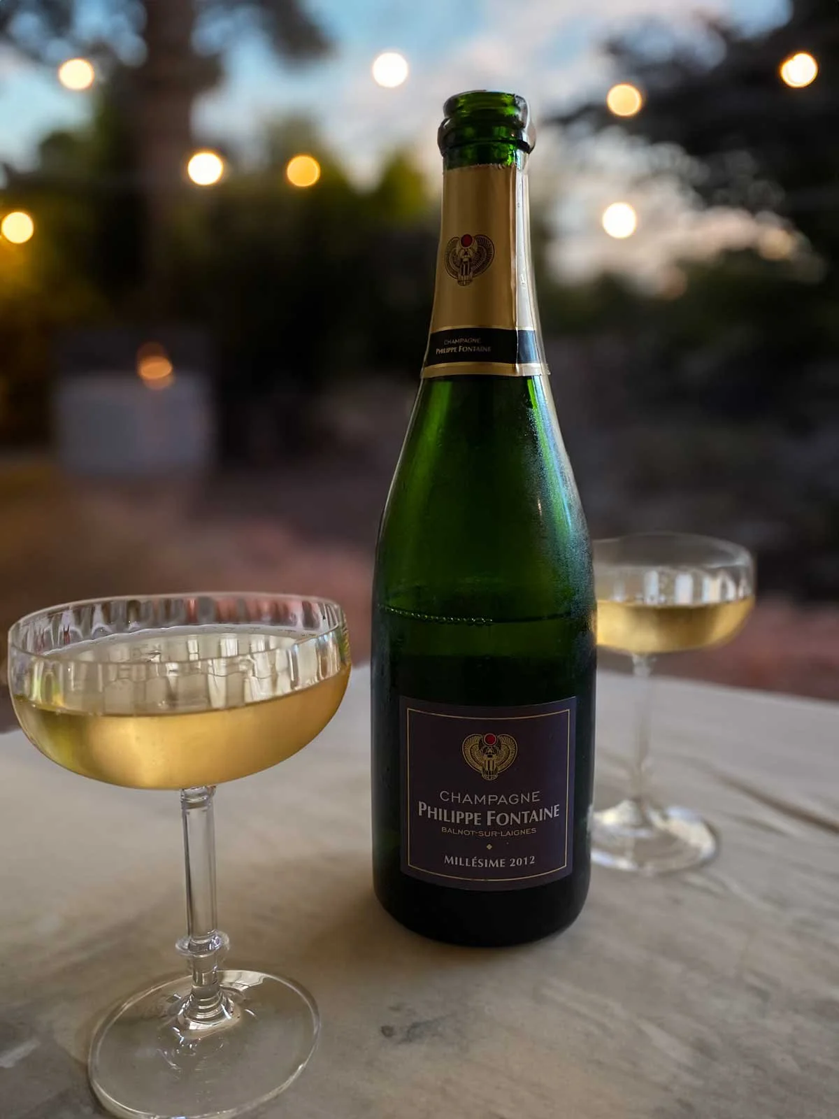 A bottle of Philippe Fontaine champagne with two filled champagne glasses on a white table, blurred outdoor background with string lights at dusk for bar service at Greenlands Farm Wedding Venue..