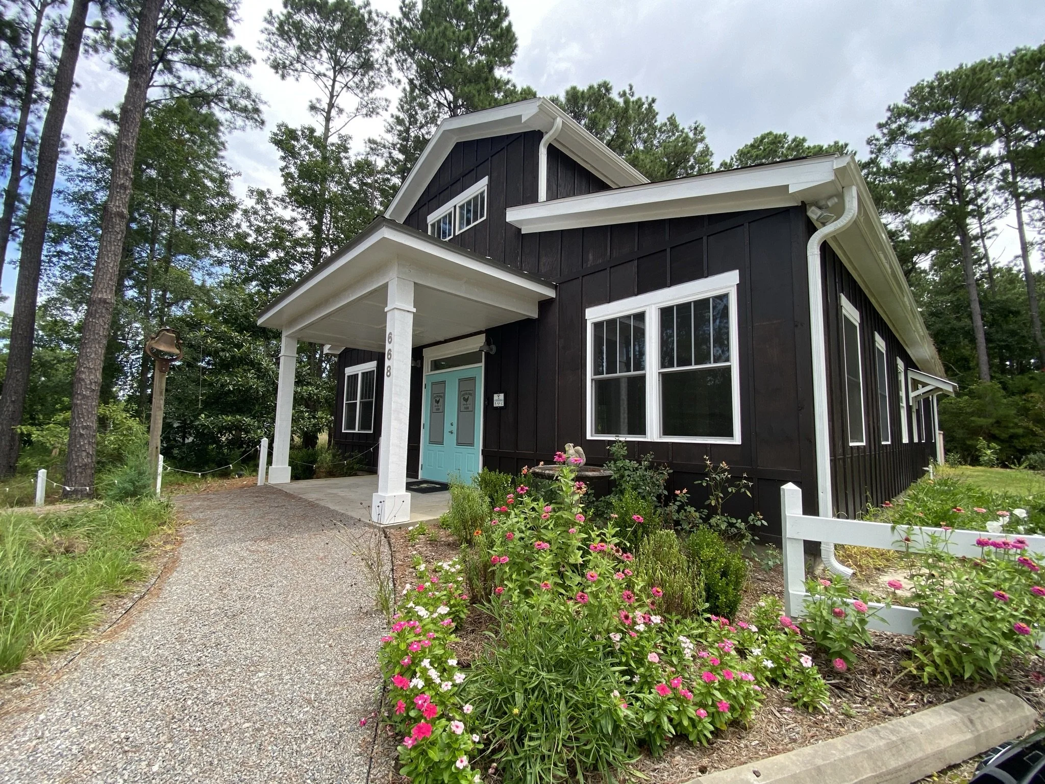 Barn style venue hall with white trim and blue front door, surrounded by green trees and blooming flowers, with a gravel pathway leading to the entrance.