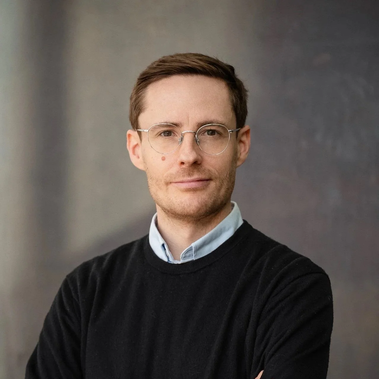 A man with short light brown hair and glasses wearing a blue suit and light blue dress shirt, posing against a plain white background.