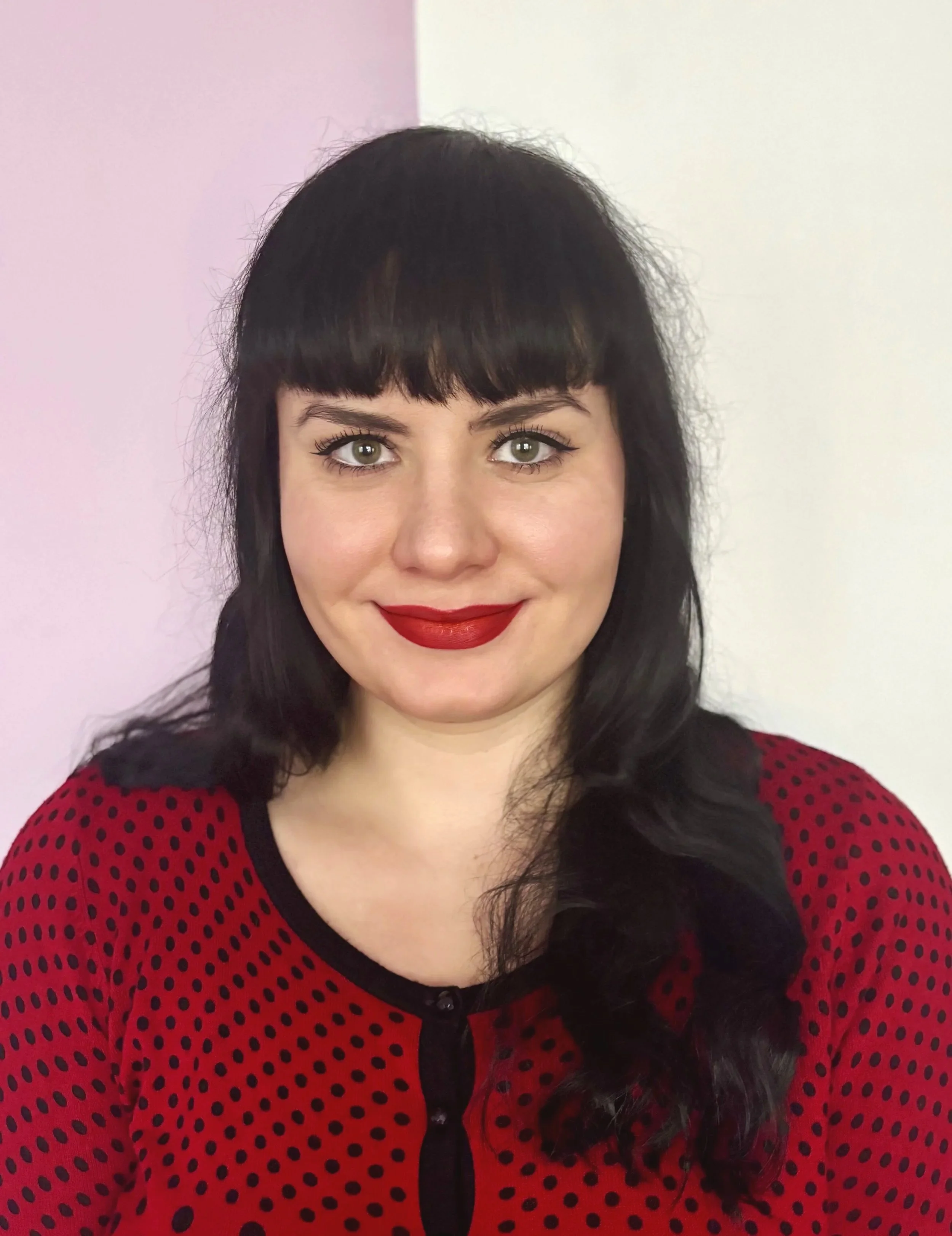 A woman with black hair and bangs wearing a black top, red lipstick, and a leopard-print and blue scarf around her neck, standing in front of a door with glass panels.