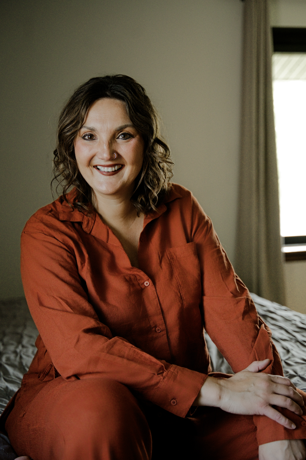 A woman with curly brown hair, wearing a rust-colored outfit, smiling while sitting on a bed in a cozy, well-lit room.