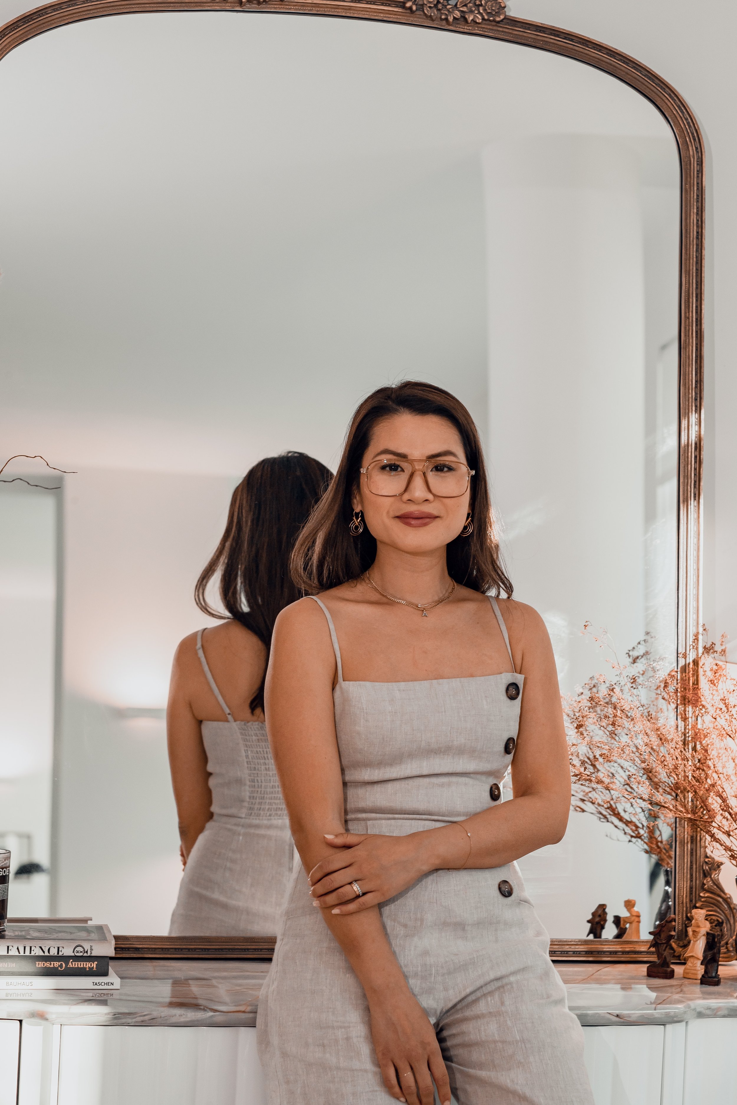 A woman wearing glasses and a beige dress with buttons, standing in front of a large mirror, looking at the camera, with a reflection of herself visible in the mirror. There are books and a decorative vase with dried flowers on a surface in front of her.