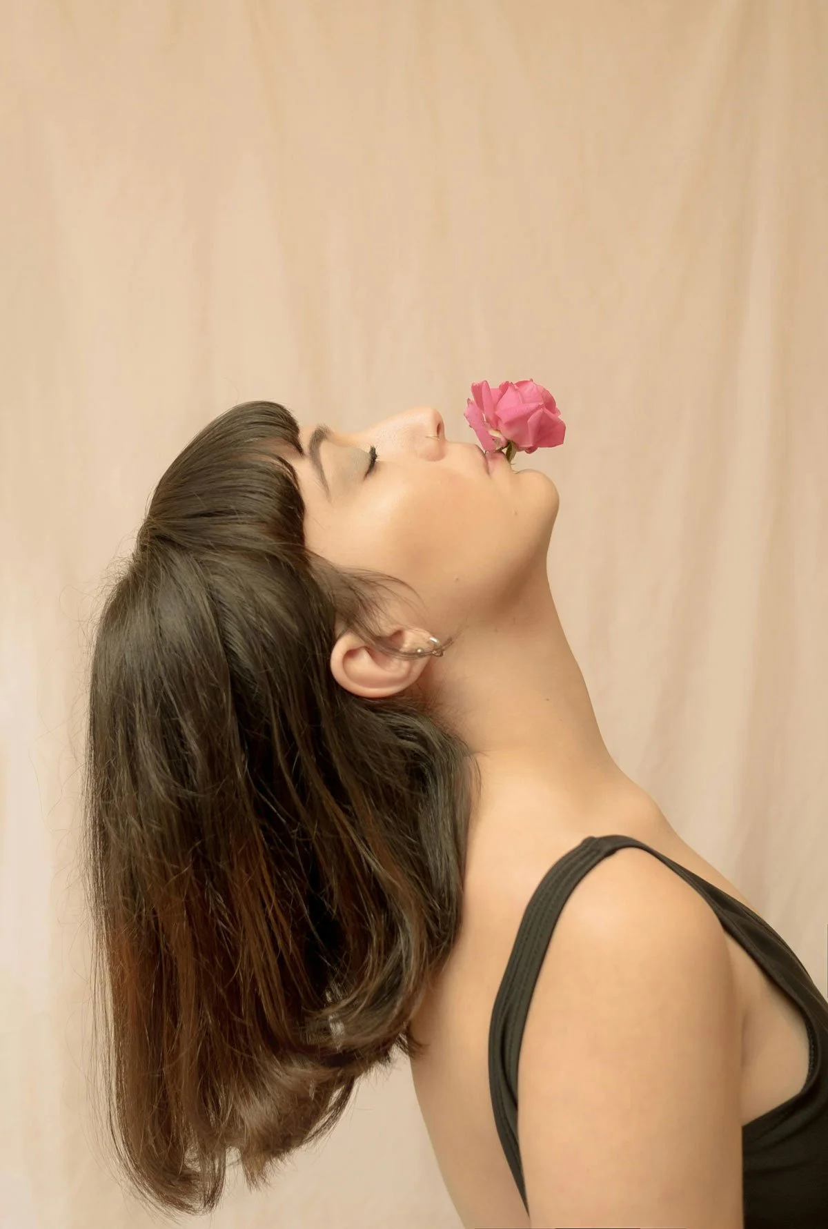 A woman with shoulder-length brown hair and a black tank top, with a pink flower near her lips, facing sideways against a neutral background.