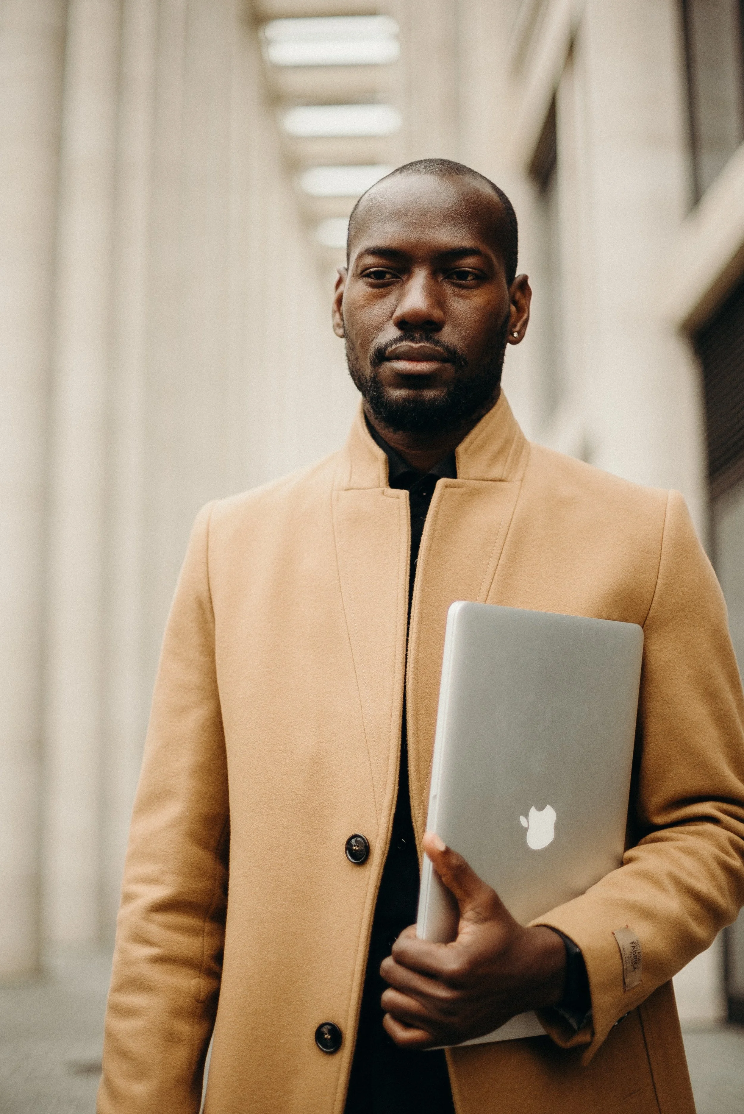 A man holding a silver laptop with a white apple logo under a covered walkway, wearing a tan coat over a black shirt.