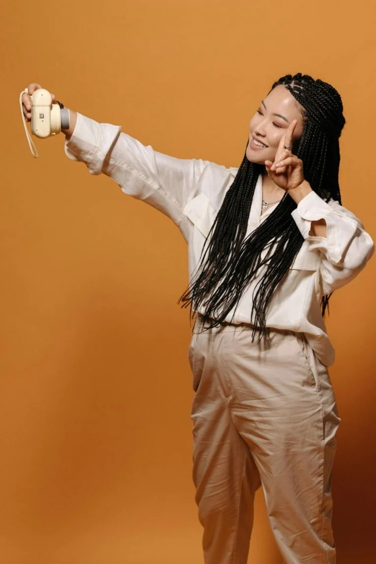 A young woman with long braided hair taking a selfie with a vintage camera against a solid orange background.