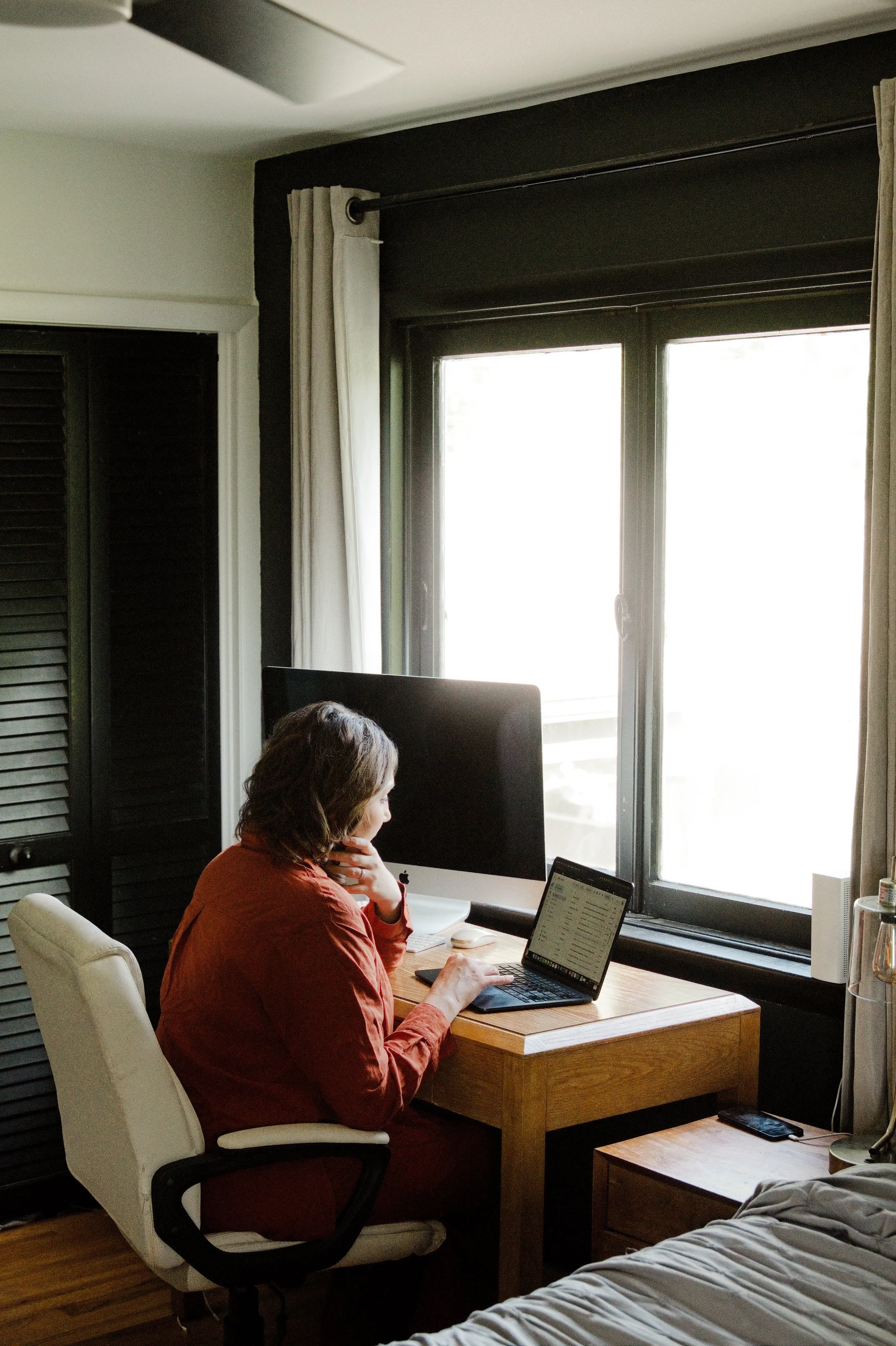 A woman sitting at a wooden desk working on a laptop in a cozy home office, with a large window to her right allowing natural light, and a bed with gray bedding visible in the foreground.