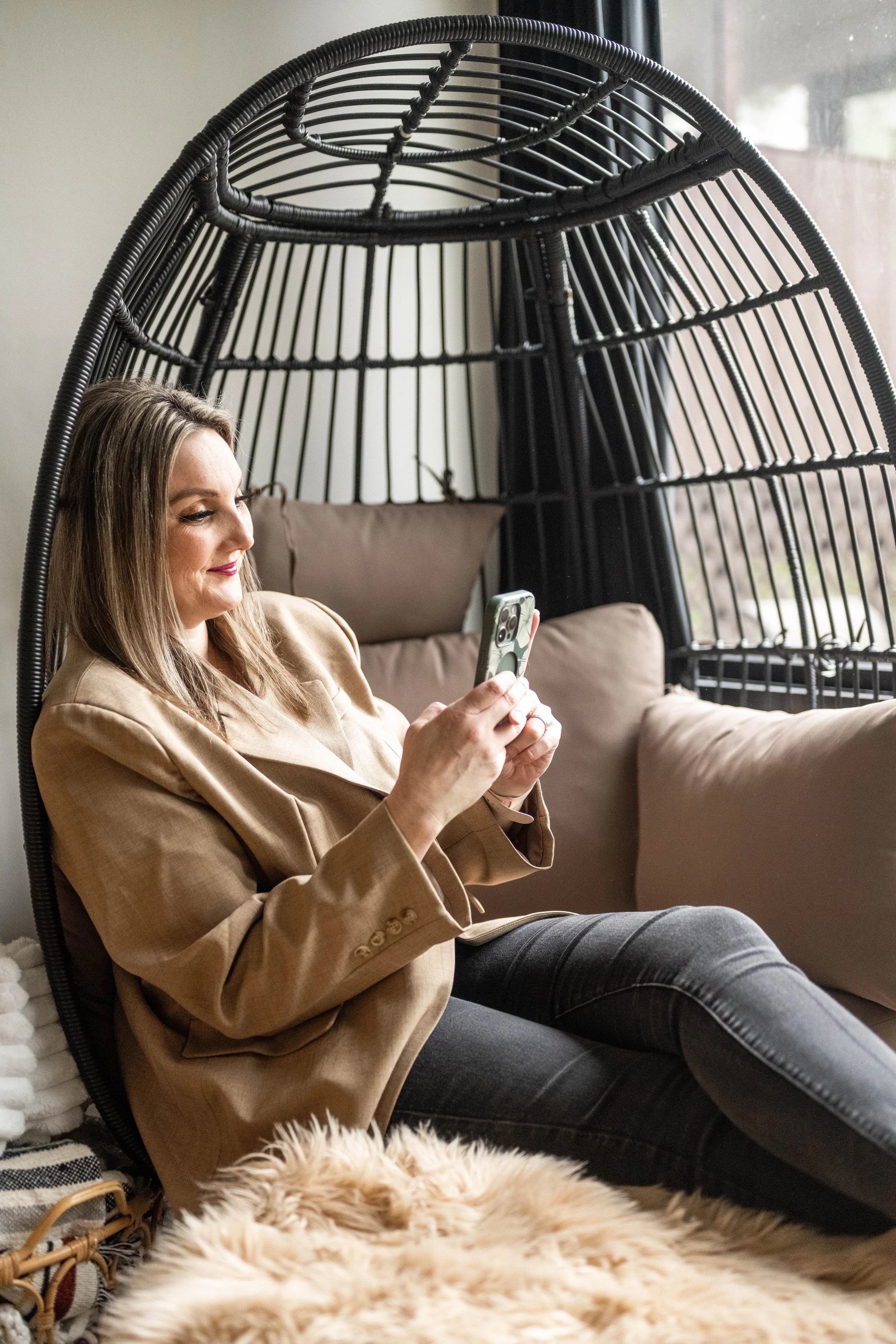 A woman with brown hair wearing a tan blazer and dark pants sitting in a black wicker egg-shaped chair, looking at her phone, in a cozy indoor setting by a window.