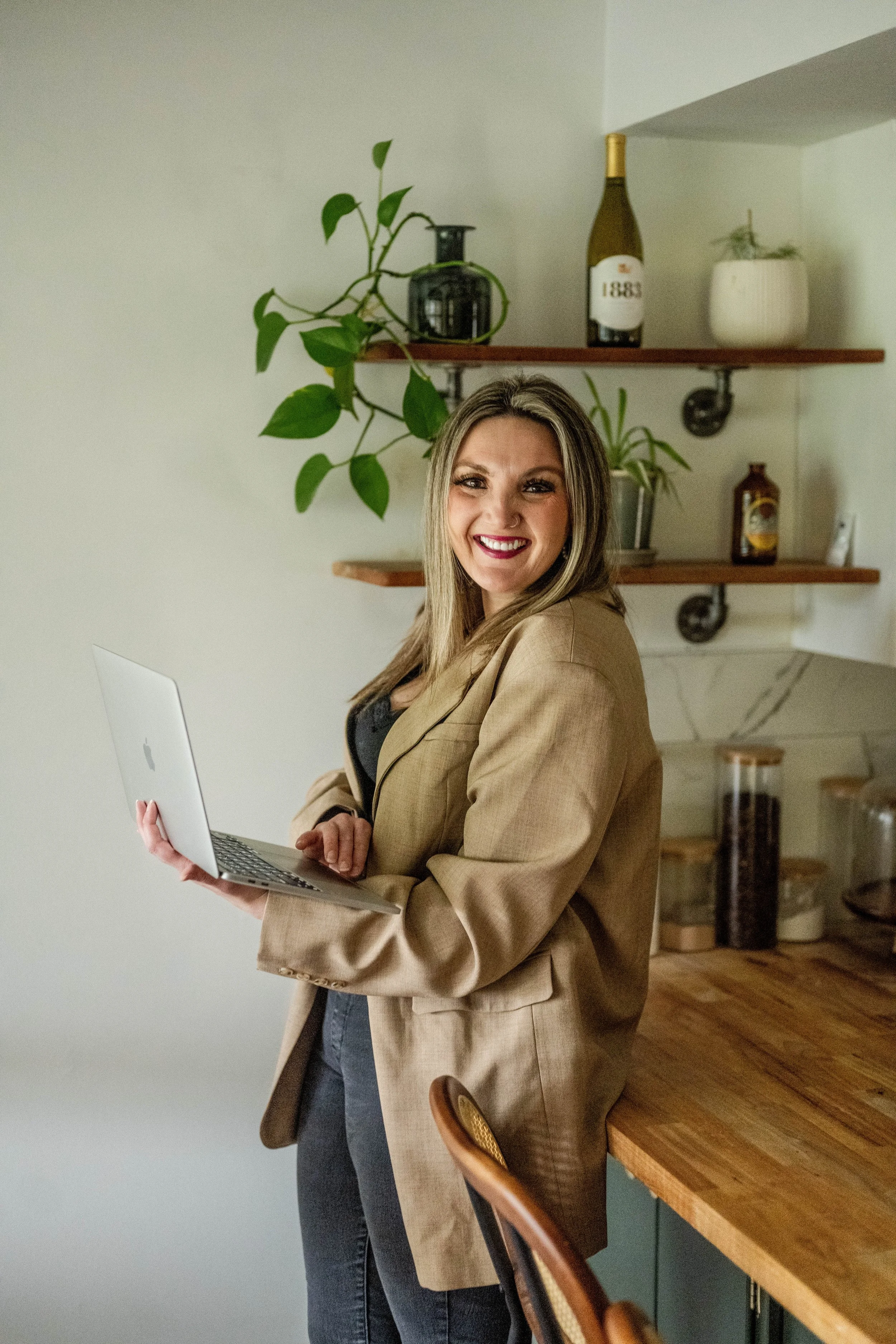 A woman smiling and holding a laptop in a kitchen with wooden shelves and cabinets, plants, and decorative items.