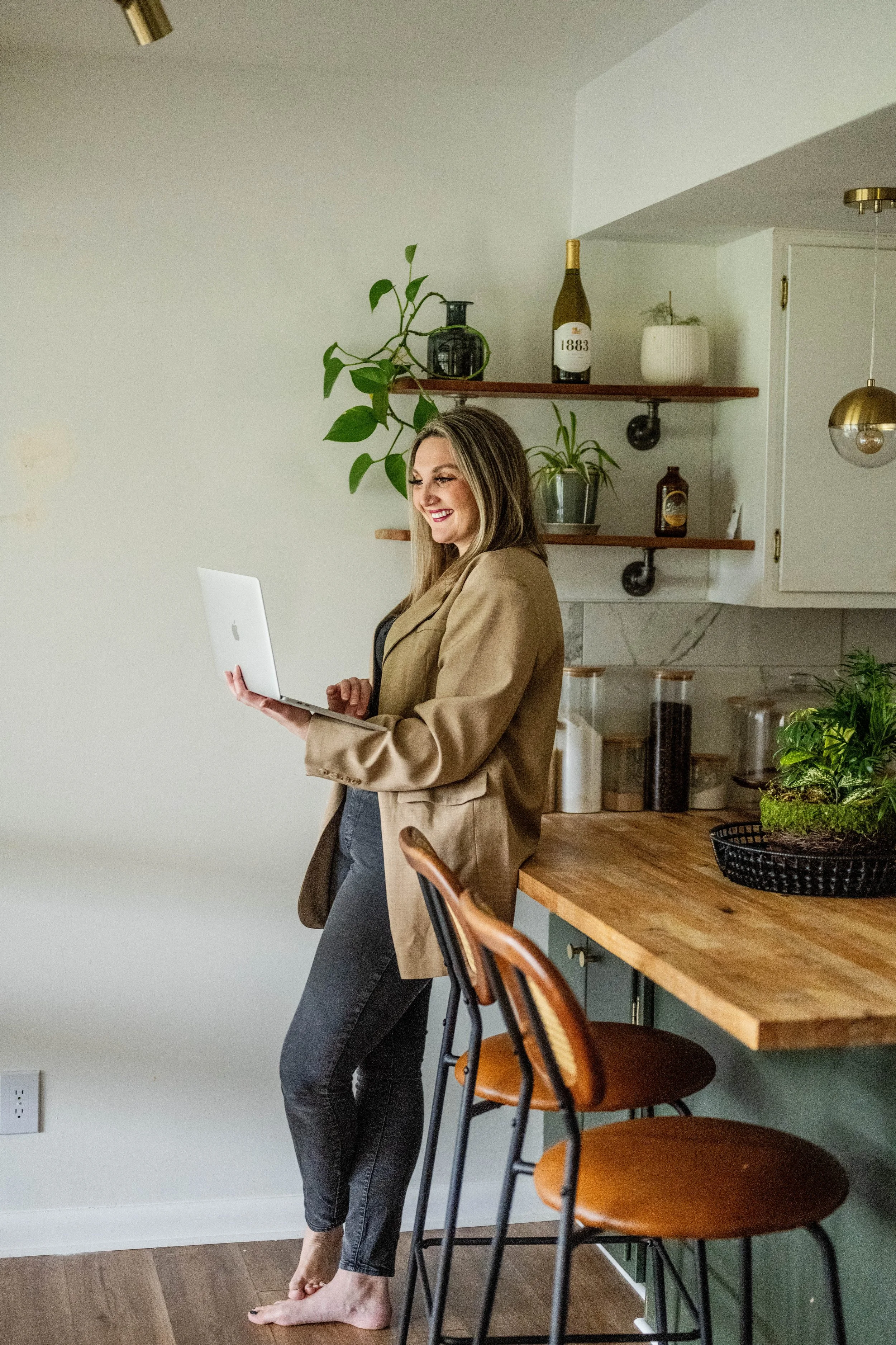 A woman standing barefoot in a kitchen, smiling while looking at her laptop, with a wooden countertop and green plants in the background.