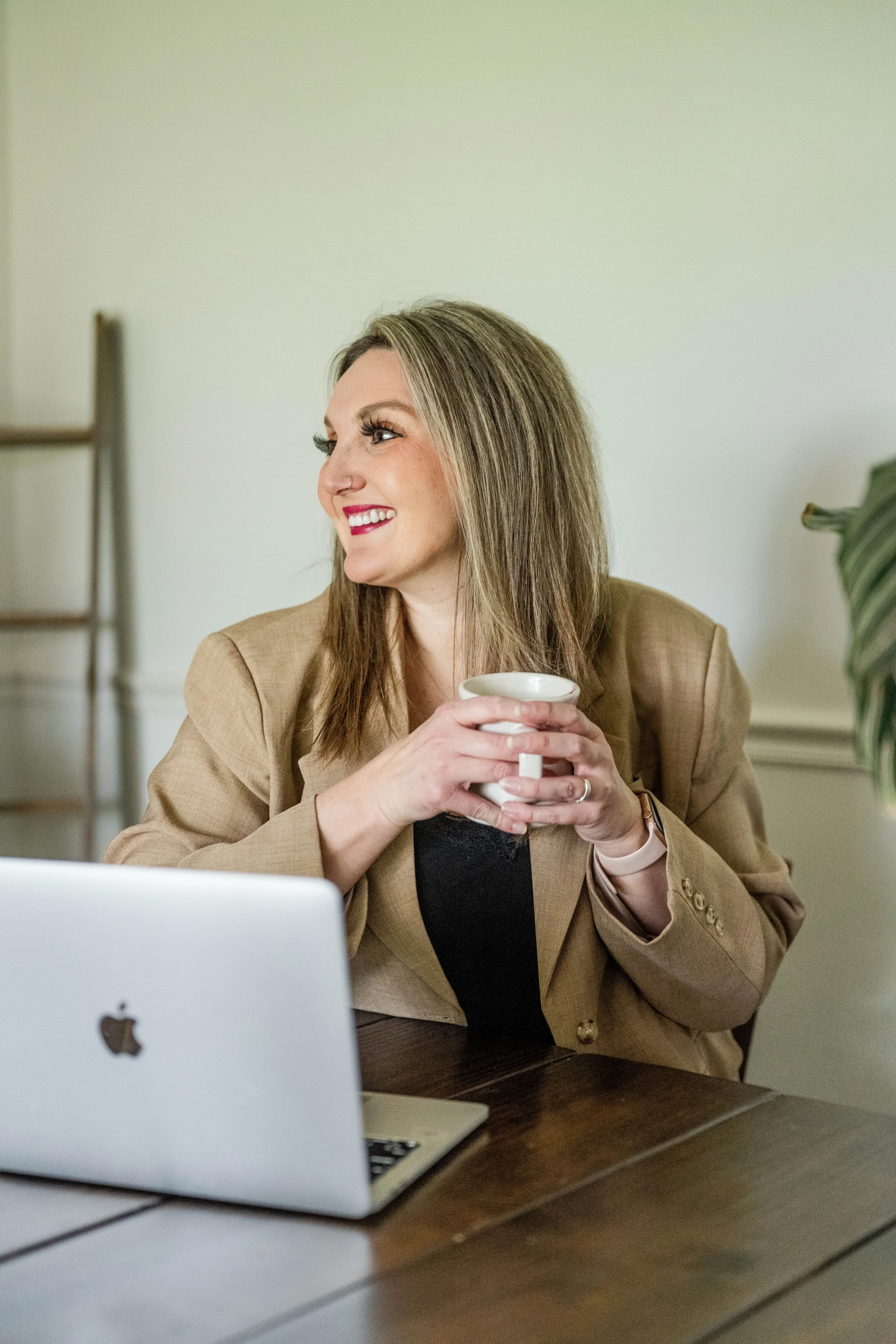 A woman with blonde hair smiling, holding a white mug, sitting at a table with a silver laptop in front of her, in a well-lit room.