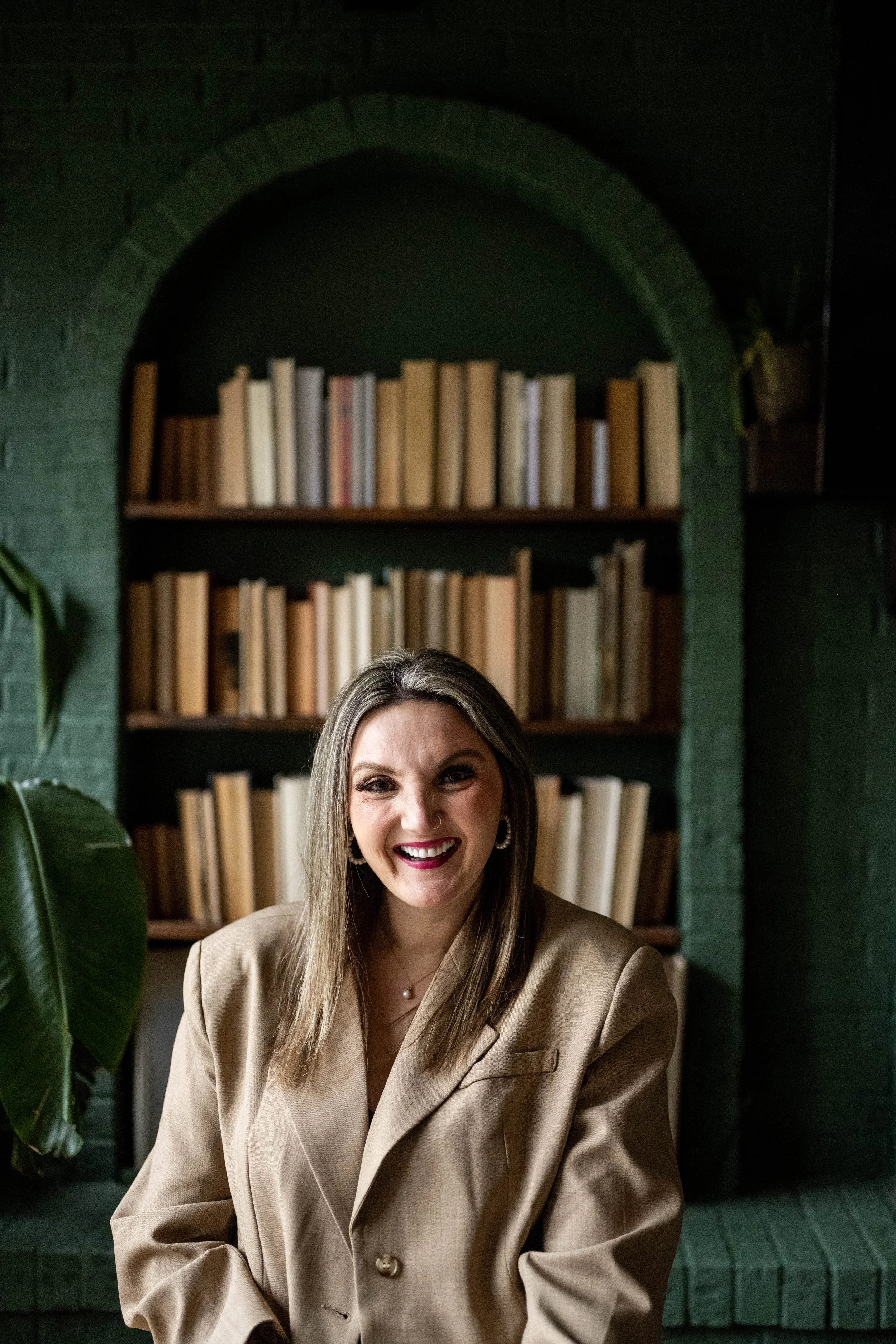 A woman with long hair smiling, wearing large hoop earrings, a beige blazer, and a necklace, seated in front of a bookshelf filled with books, inside a room with green walls and a brick arch.