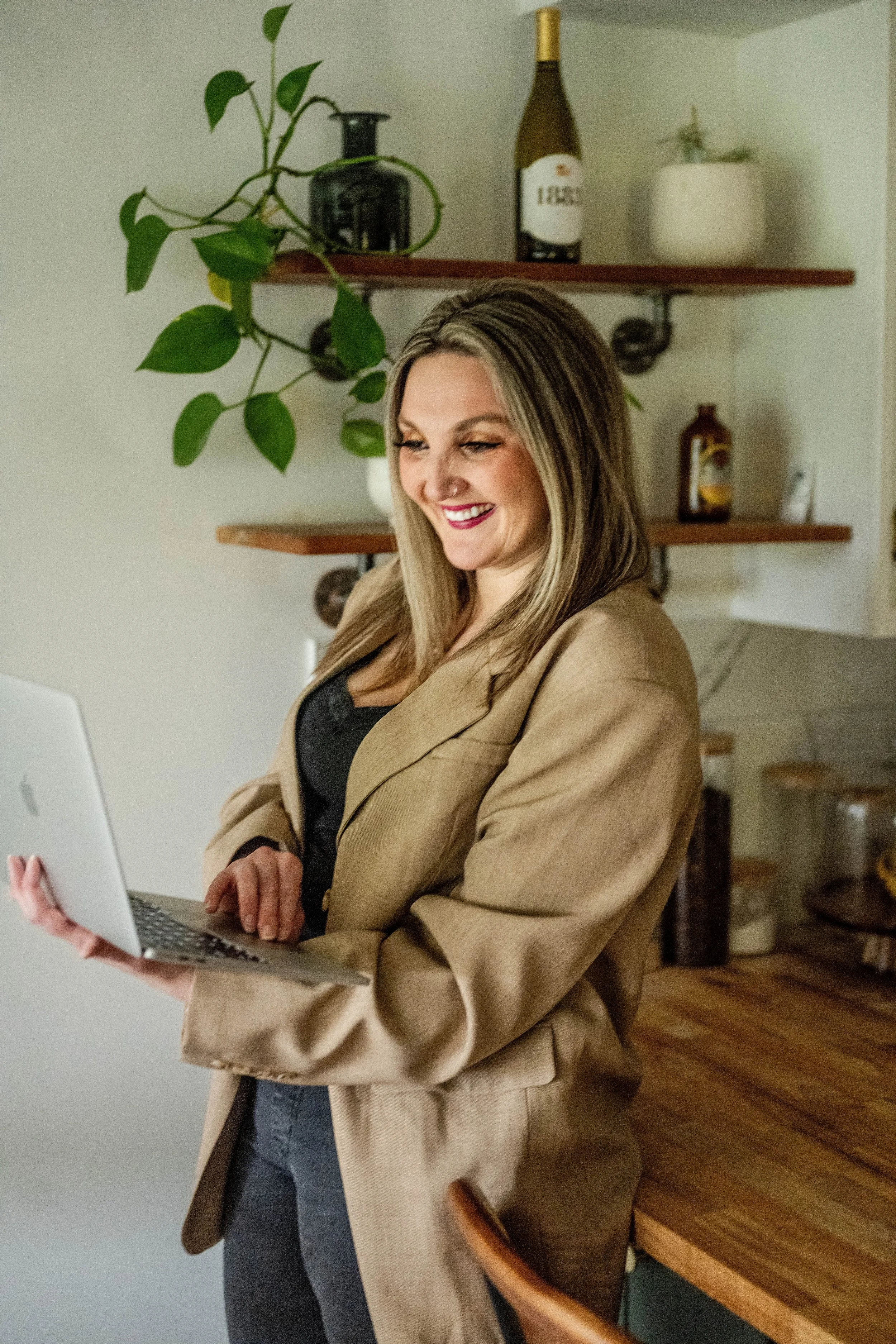A woman with blonde hair and a beige blazer smiling while looking at a silver laptop in a cozy home kitchen with wooden shelves and various bottles and plants.