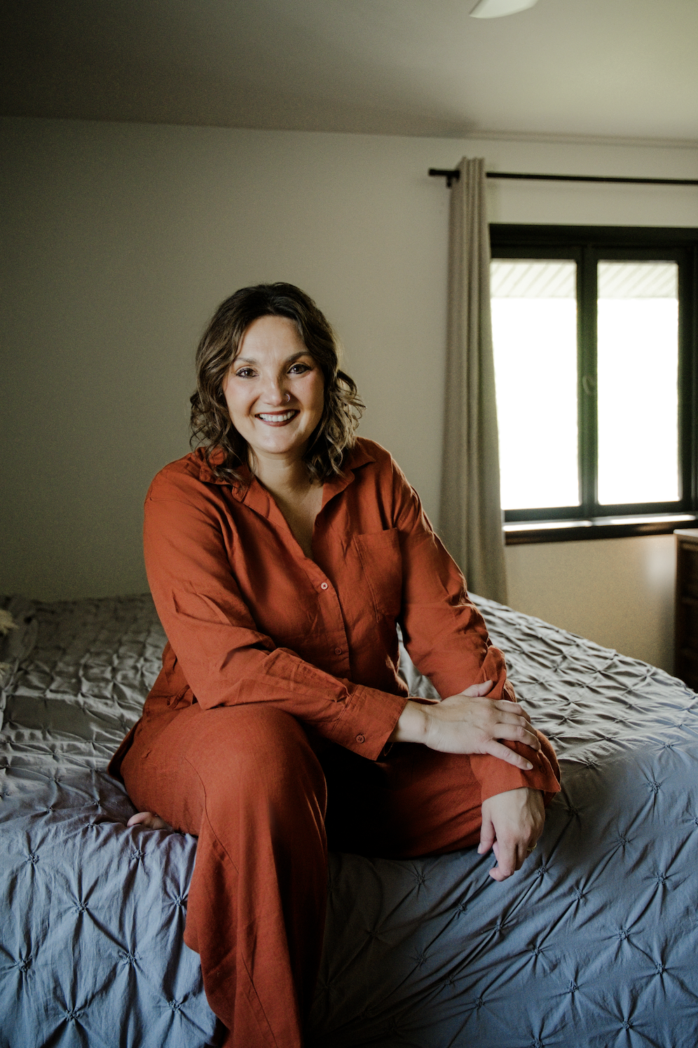 A woman with curly brown hair, wearing an orange pajama set, sitting on a bed in a bedroom with a large window in the background.