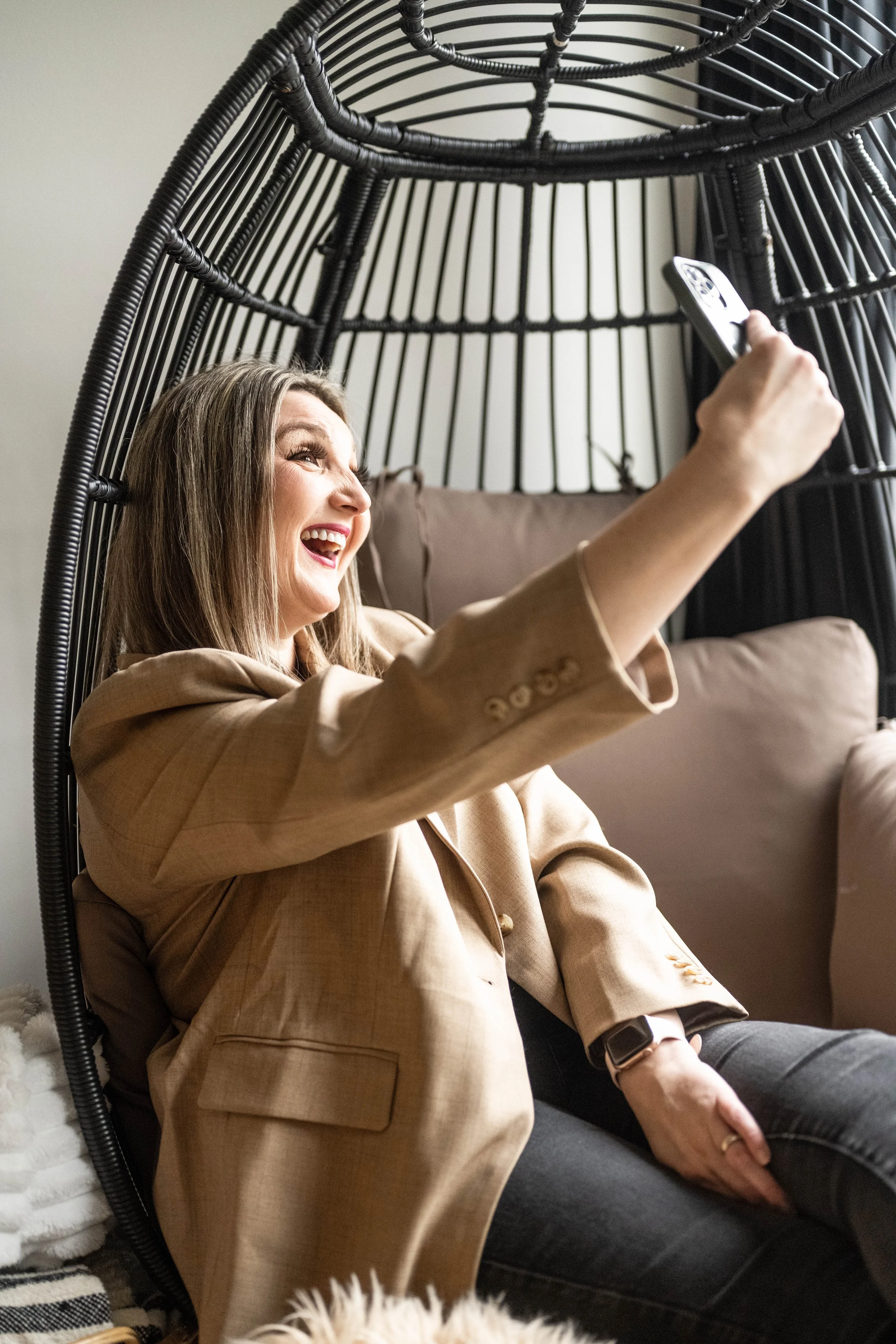 A woman sitting in a black, woven, hanging chair, smiling and taking a selfie with her smartphone.