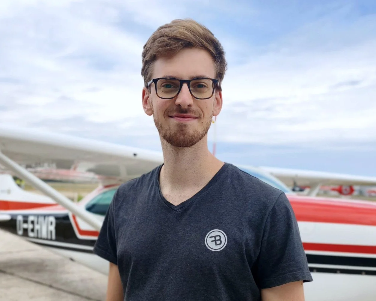 Fluglehrervorstellung Christopher Lutz: Ein junger Mann mit Brille, kurzen braunen Haaren und Bart trägt ein graues Polo-Shirt mit einem FSB-Logo auf der linken Brustseite, steht vor einer grauen Wand mit großen weißen Buchstaben.