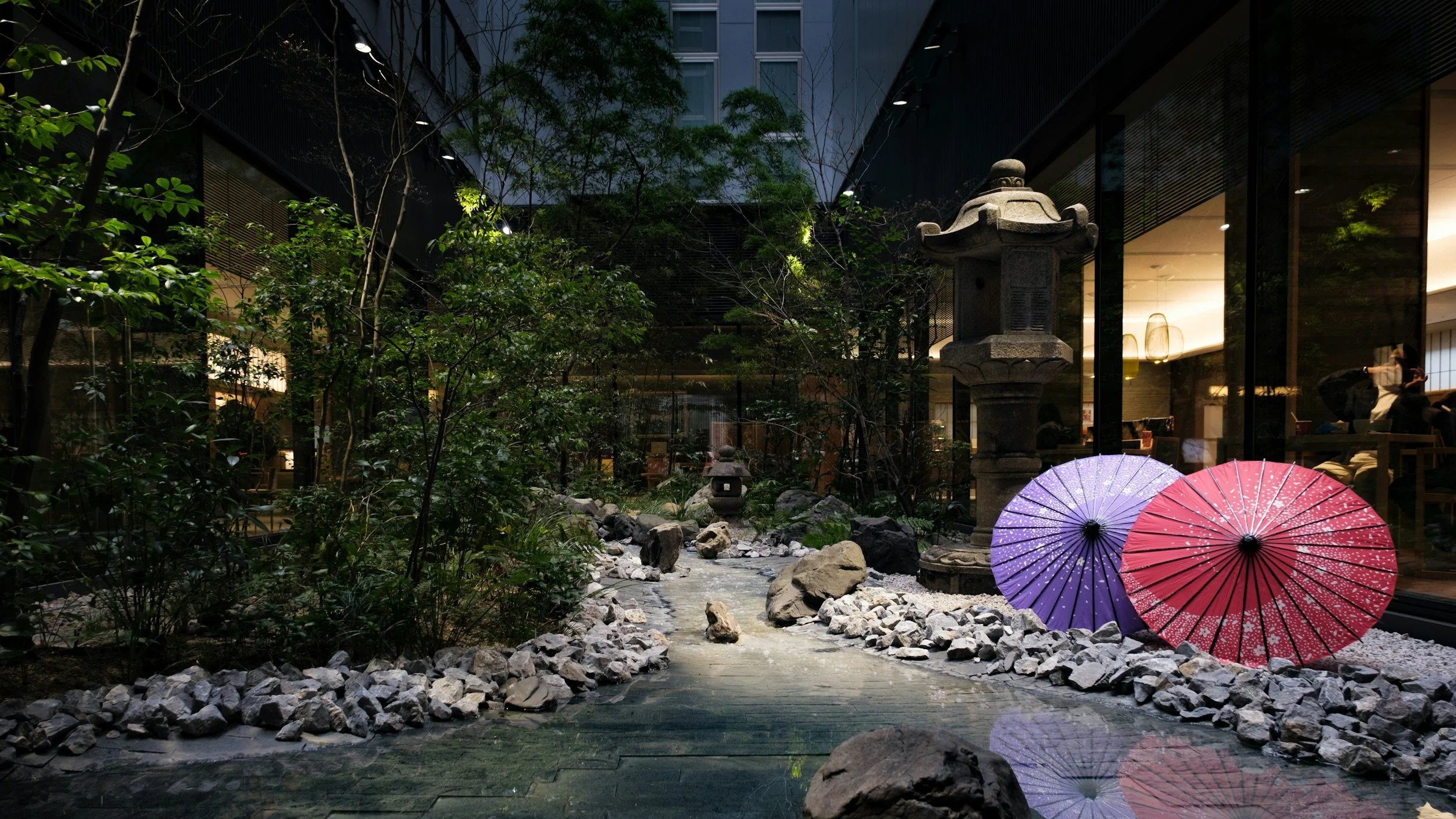 A serene Japanese indoor garden featuring a stone lantern, rock stream, and traditional red and purple umbrellas, illustrating the blend of tradition and modernity in Tokyo business spaces.