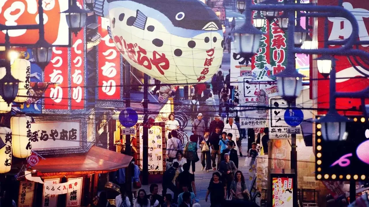 A crowded street scene in Japan with many brightly lit signs and lanterns, including a large inflatable fugu (pufferfish) sign, symbolizing the vibrant Japanese service industry and the consumption conundrum.