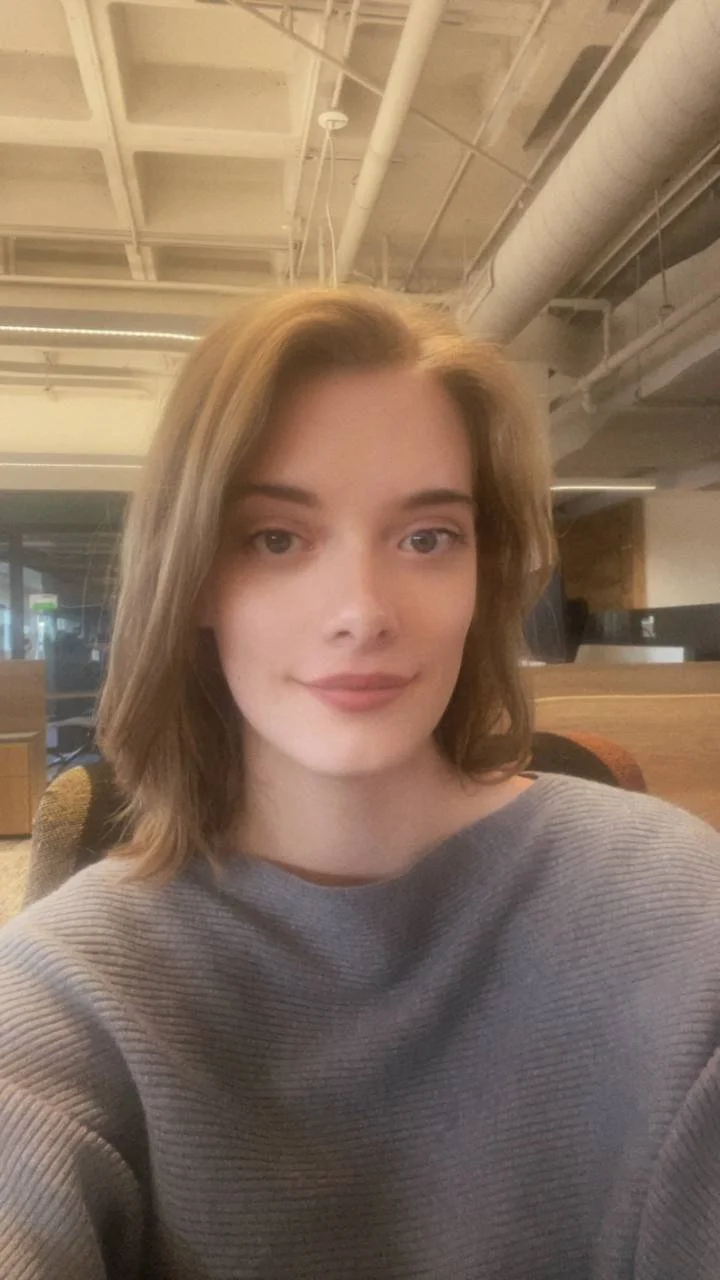 young woman in a grey sweater smiling at the camera in a building with an exposed industrial ceiling