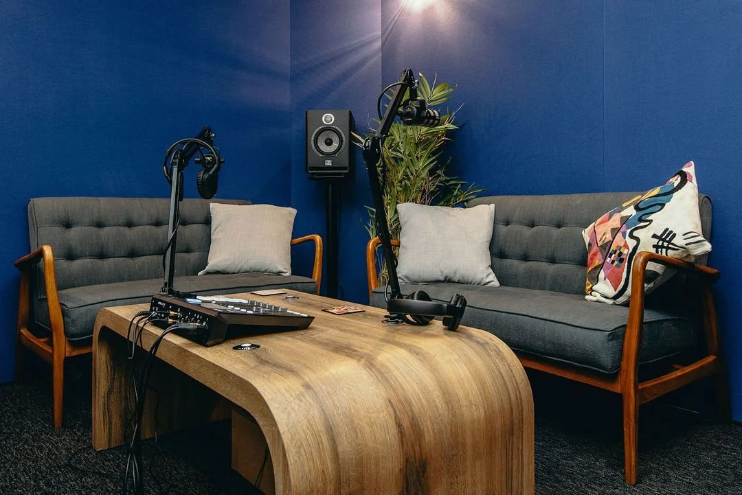 Podcast recording setup with two sofas, microphones, audio equipment on a wooden table, and a speaker mounted on a blue soundproof wall, creating a cozy and professional recording environment.