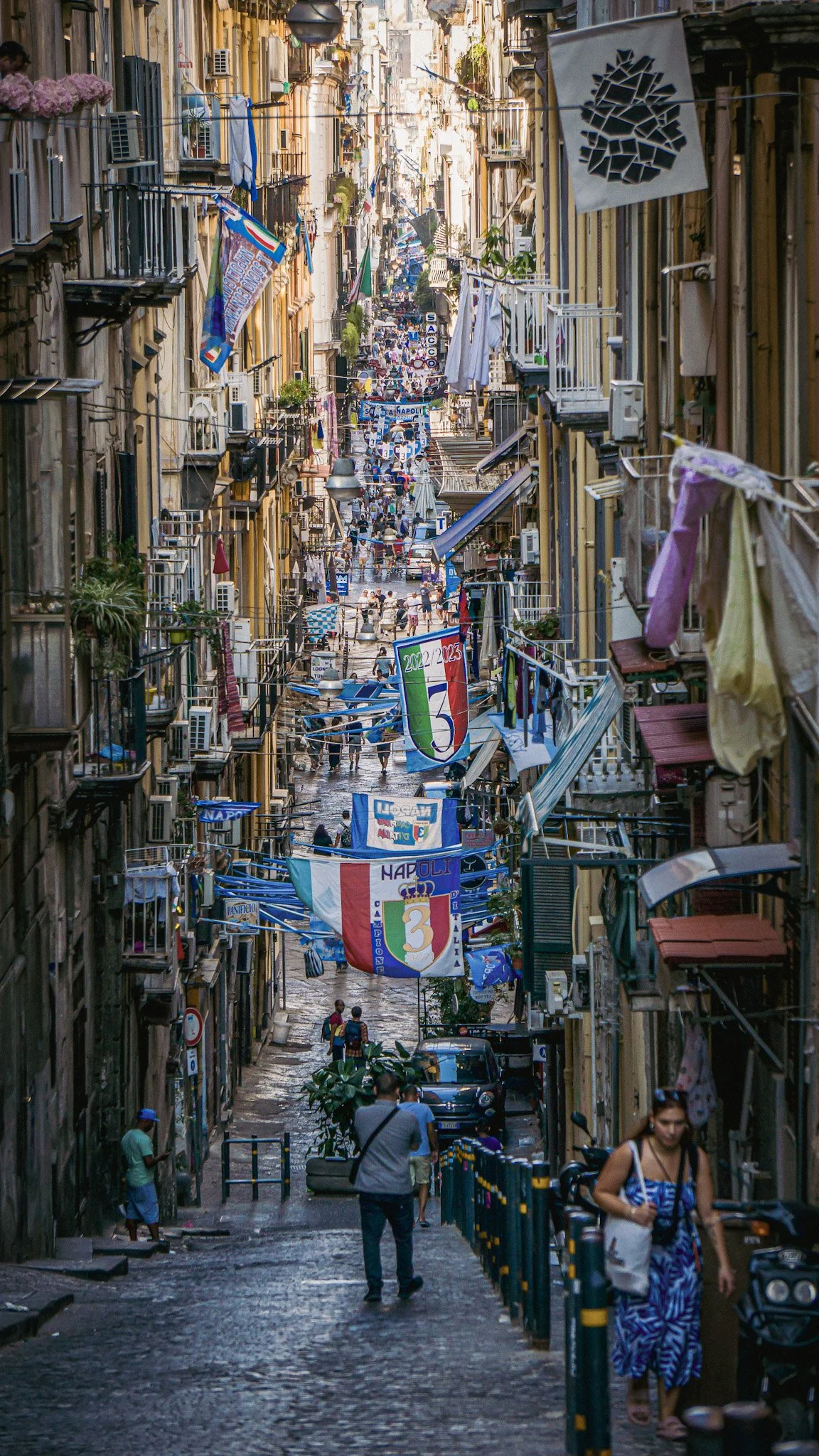 A narrow street in Naples, Italy, decorated with blue, white, and red banners and flags, crowded with people, shops, and residential apartments.