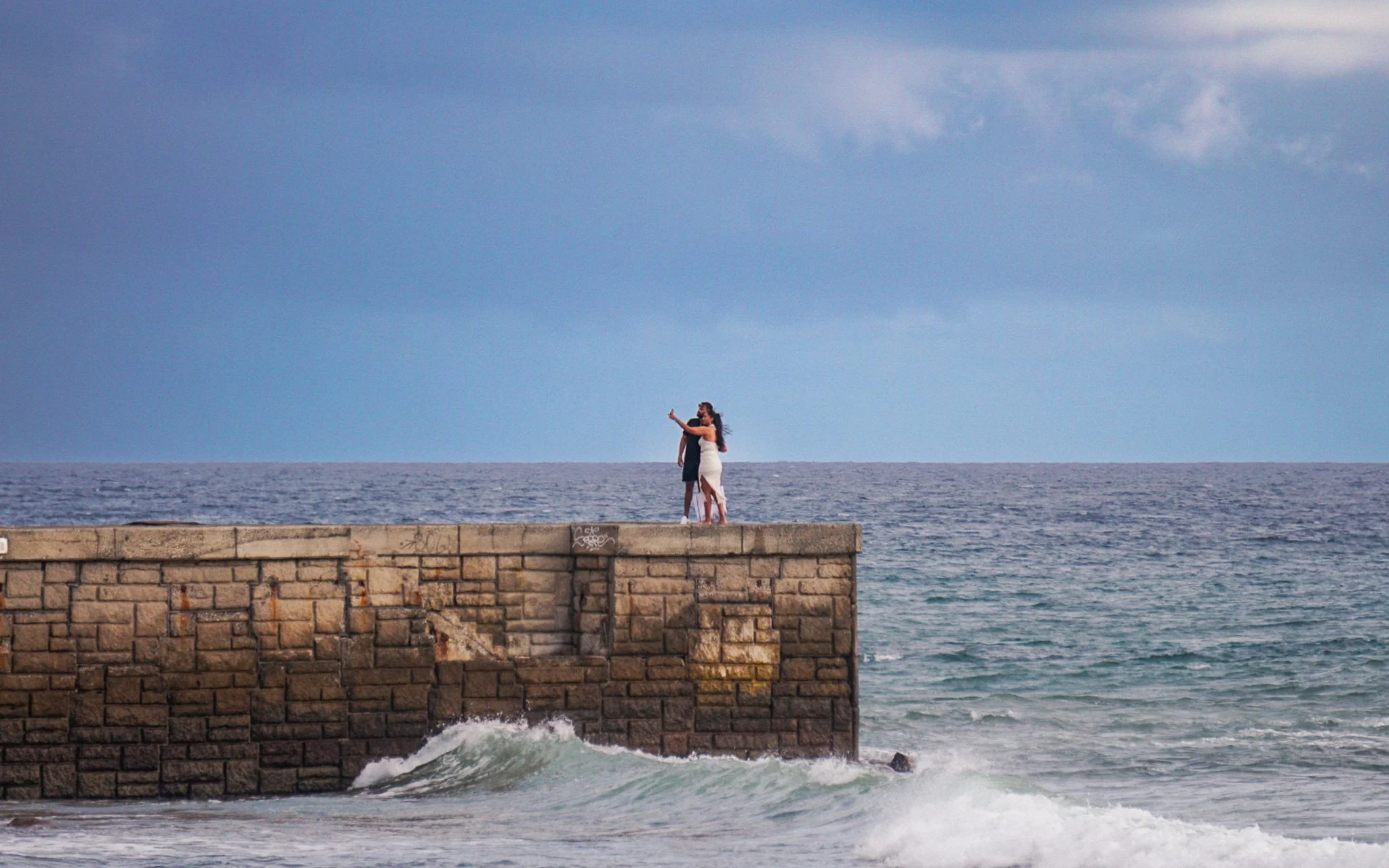 A woman in a white dress and a man in dark clothing stand on a stone pier, with the ocean behind them and waves at the base of the pier, under a blue sky with some clouds.