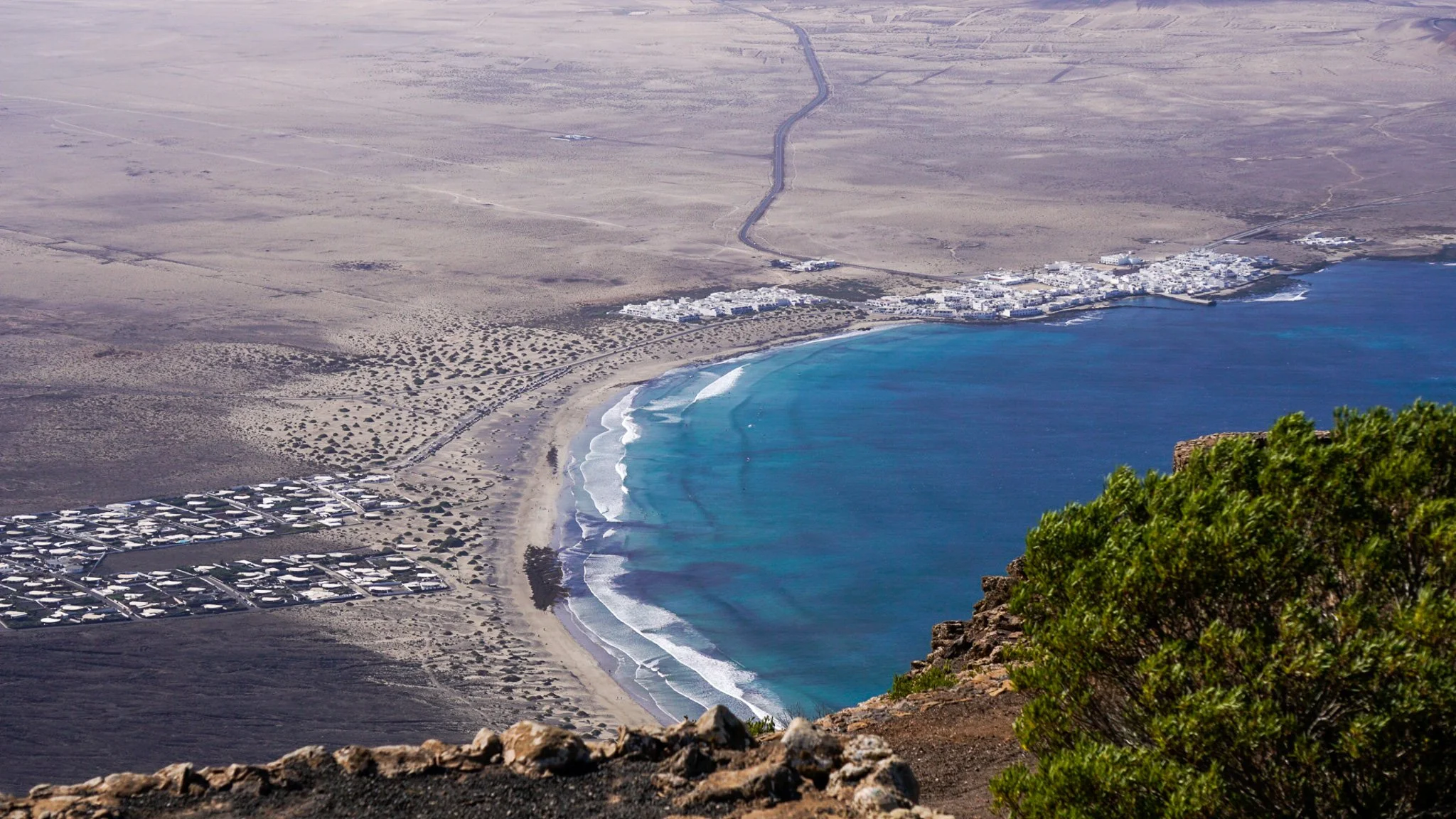 Aerial view of a coastal town with white buildings, sandy beach, and turquoise ocean, surrounded by arid desert landscape, with some green bushes in the foreground.