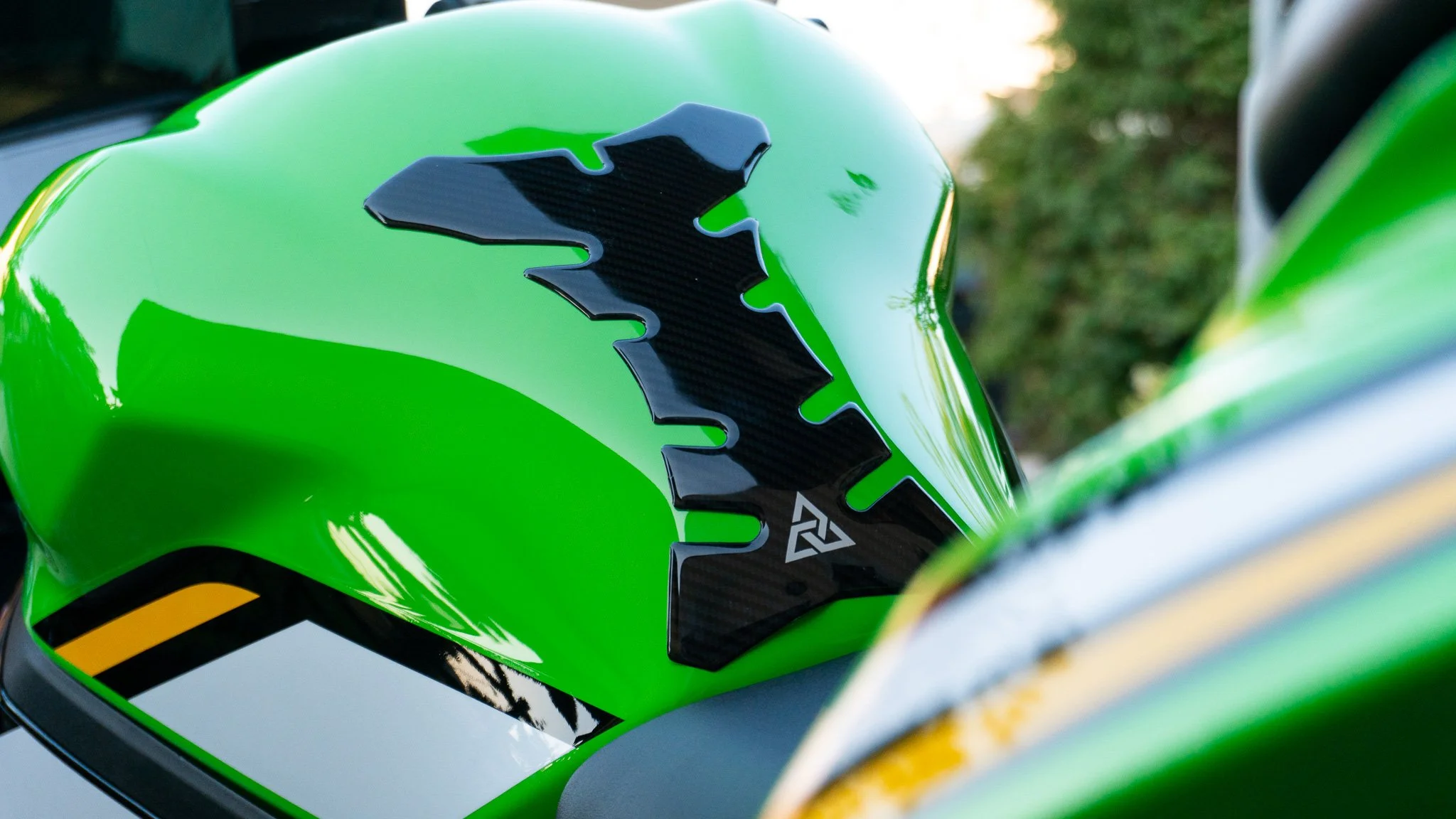 Close-up of a bright green motorcycle gas tank with a black carbon fiber decal and graphic design, with a blurred background of trees and sky.