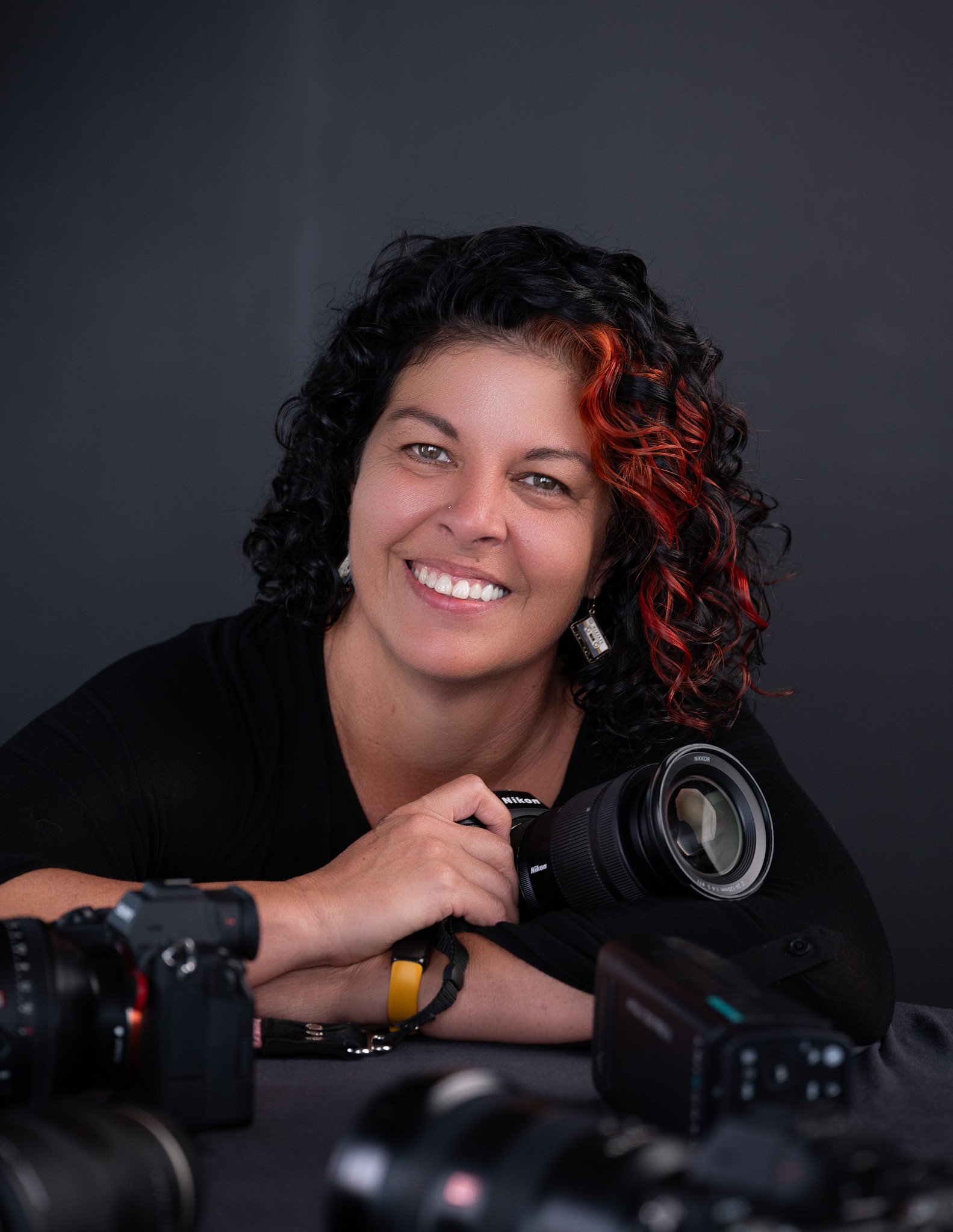Person with curly hair holding a large camera lens, wearing a yellow sweater and black polka dot shirt against a pink background.