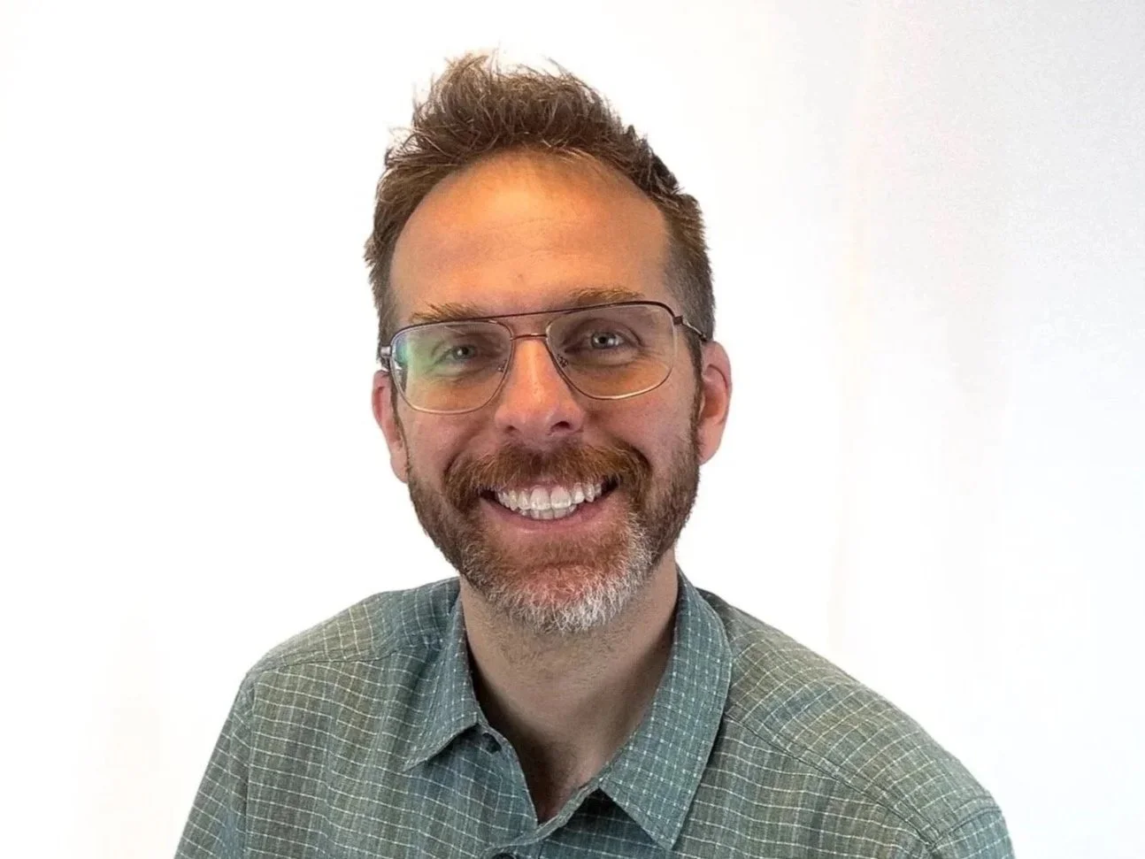 A smiling man with glasses, a beard, and short, styled hair, wearing a checkered shirt, standing against a plain white background.