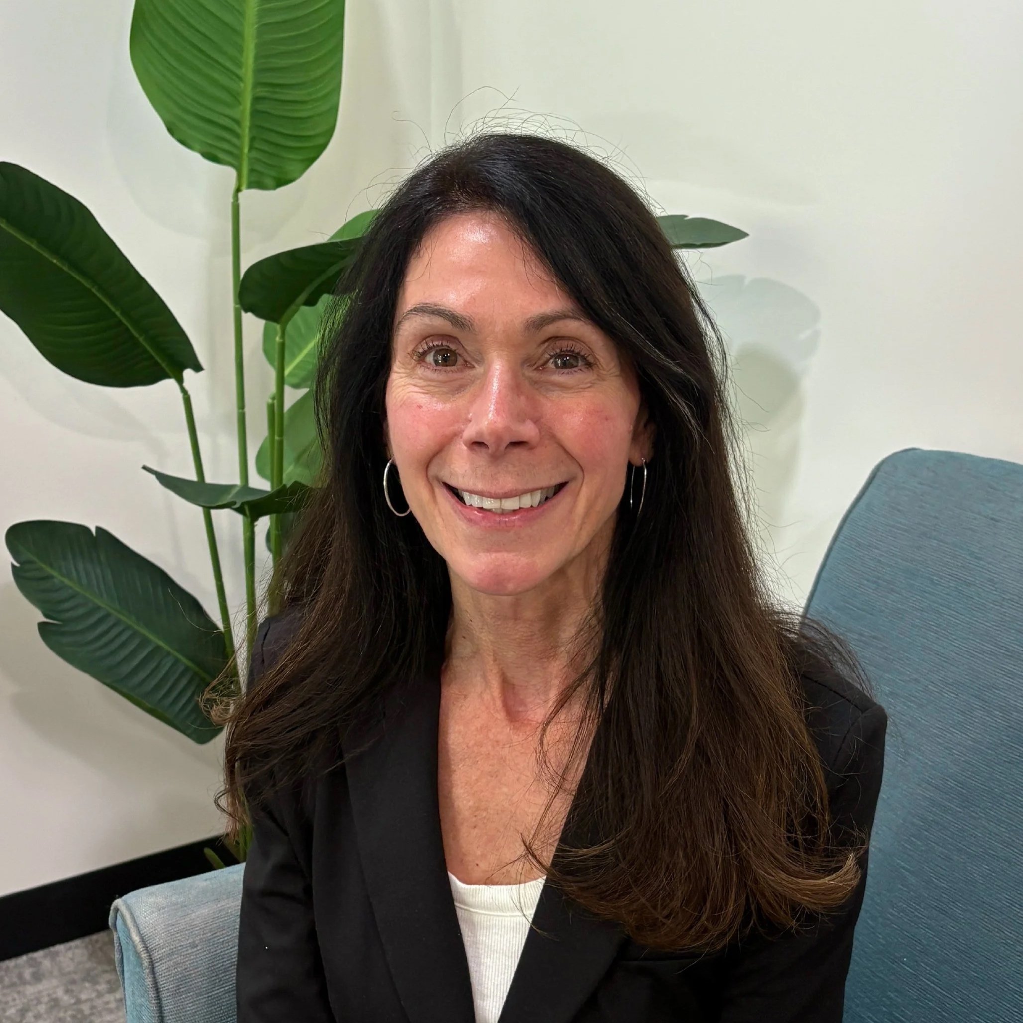 Close-up of a smiling woman with long dark hair, sitting on a gray office chair, with large green houseplant leaves behind her and a light-colored wall in the background.