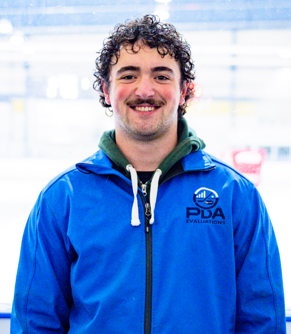 Young man with curly dark hair and a beard wearing a blue jacket with PDA Evaluations logo, smiling in front of a plain wall.