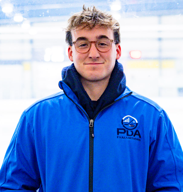 Young man with light brown hair, wearing a black suit with a subtle checkered pattern, a white shirt, and a black tie, standing against a plain light background.