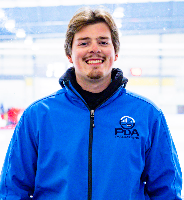 A young man with shoulder-length light brown hair and a goatee smiling, wearing a blue jacket with the logo 'PDA EVALUATIONS' on it, standing inside an ice rink.