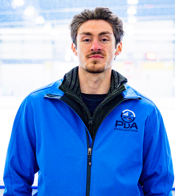 Young man with brown hair and beard wearing a blue jacket with a logo that reads 'PDA Evaluations' in an ice rink.