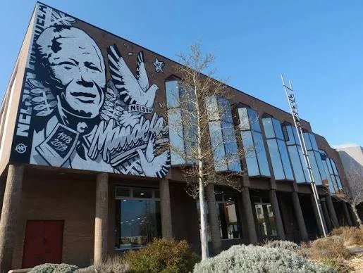A building with a large mural of Nelson Mandela on the exterior wall, featuring a dove and the words "Nelson Mandela" with a blue sky background.