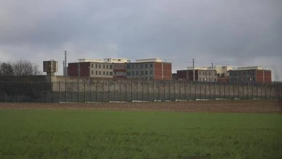 A row of multi-story buildings seen behind a fence, with a green grassy area in the foreground and a cloudy sky above.