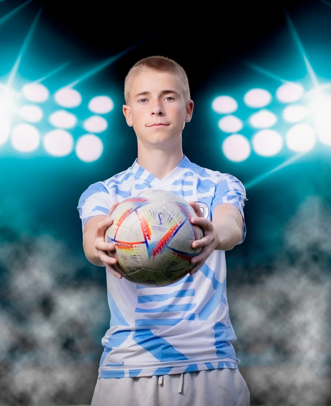 A young male soccer player in a light blue and white uniform holding a soccer ball with a stadium background and bright stadium lights.