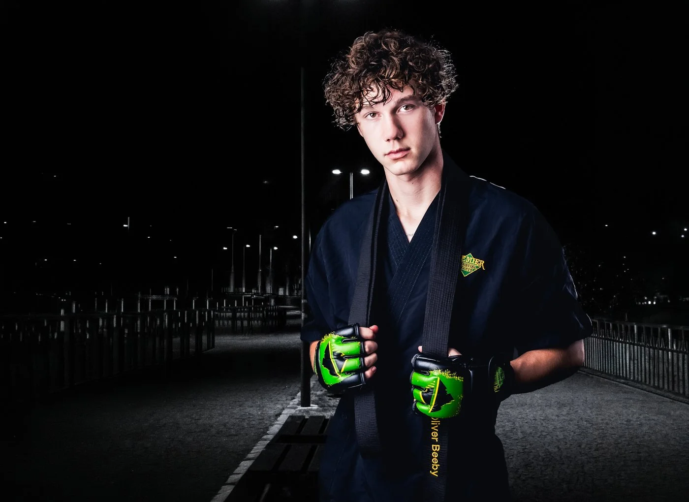 Young man with curly hair dressed in a black martial arts uniform, wearing green and black gloves, standing outdoors at night with a black background and some lights in the distance.
