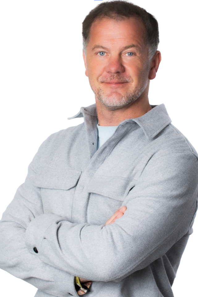 Stuart Beeby is the founder of Schoolypics , smiling slightly, wearing a light gray shirt with its collar popped, standing with arms crossed against a white background.