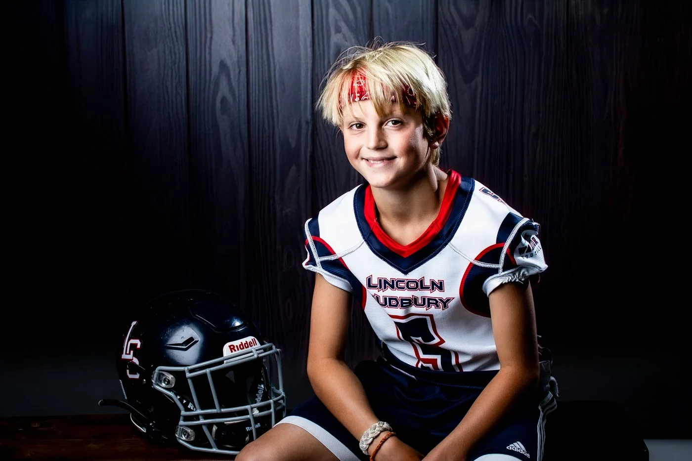 A young boy in a football uniform sitting next to a black football helmet on a wooden surface, smiling at the camera against a dark background.