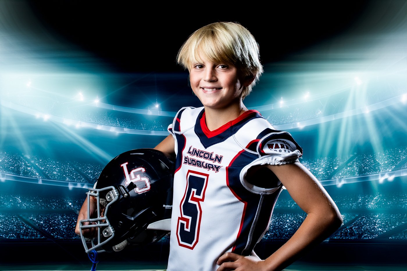 Young boy in football uniform holding a helmet, smiling, in a stadium background.