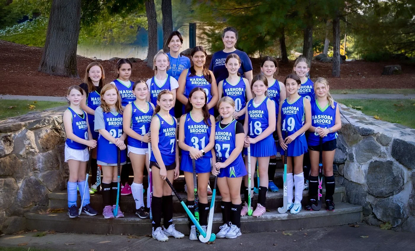 A girls' field hockey team posing outdoors on stone steps, wearing blue and black uniforms with their coach.